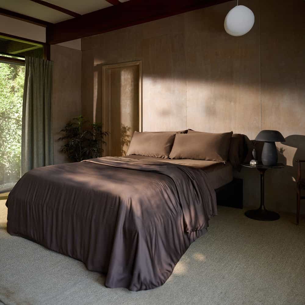 A serene bedroom with a large bed adorned with Cozy Earth's Bamboo Duvet Cover in a smooth, chocolate-brown color. The room features a neutral color palette, a nightstand with a dark lamp, a lush potted plant in the corner, curtains, and soft natural light filtering in from the window. |Color:Coffee