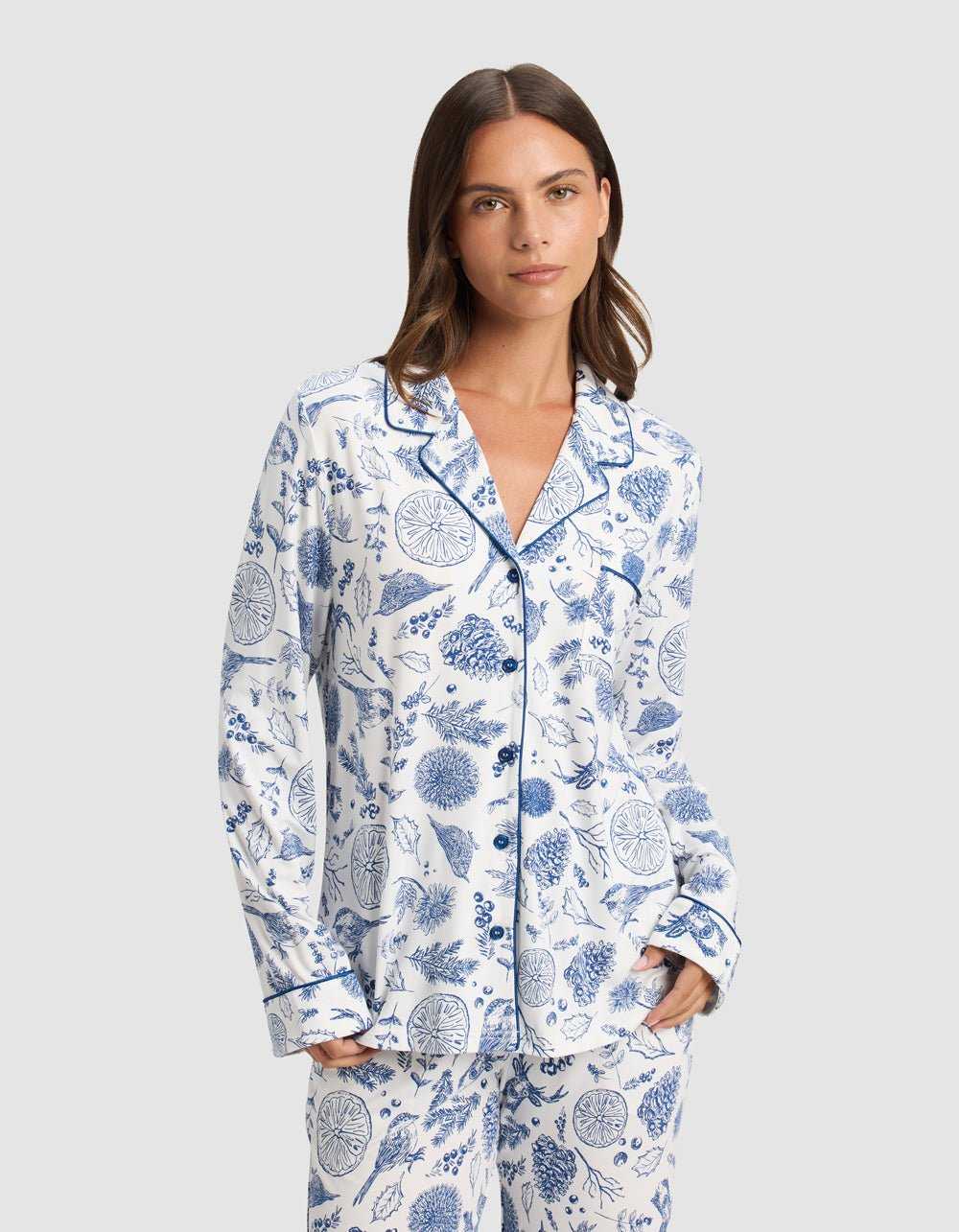 A woman with straight brown hair wears the Cozy Earth Women's Bamboo Stretch-Knit Long Sleeve Pajama Top, featuring a notched collar and blue botanical prints, standing with her hands in her pockets against a plain light background.|Color:Winter Toile Pacific