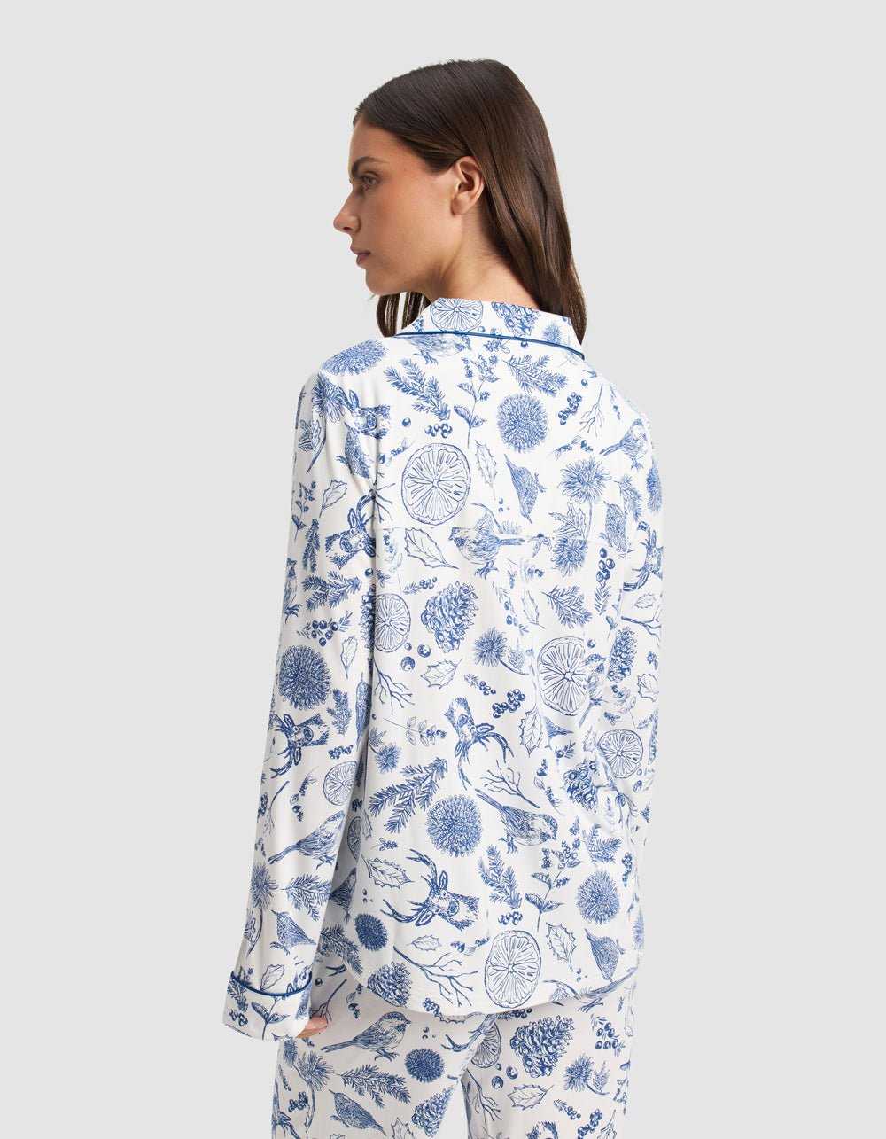 A woman with straight brown hair models the Cozy Earth Women's Bamboo Stretch-Knit Long Sleeve Pajama Top in white with a blue botanical print, standing with her back to the camera against a plain light gray background.|Color:Winter Toile Pacific