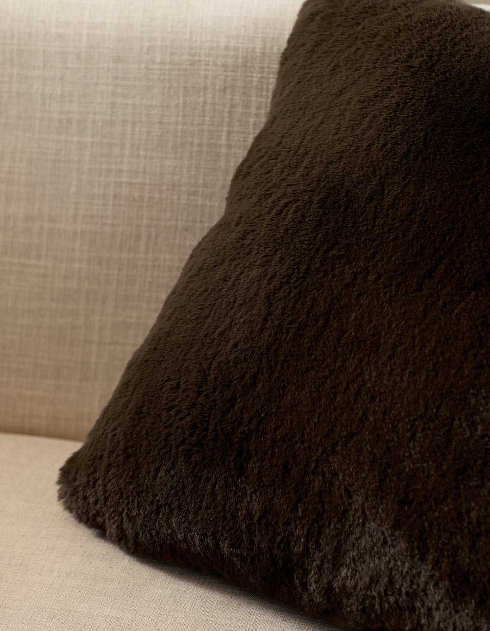 A Cozy Earth Cuddle Pillow in dark brown rests against the back of a light beige, textured fabric sofa.|Color:Espresso