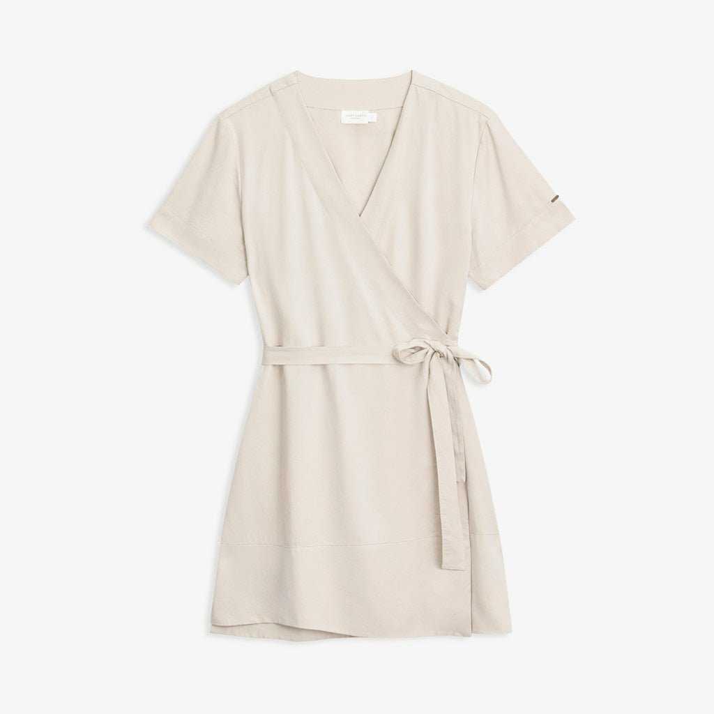 The Women's Cozy Earth Coastal Comfort Wrap Dress by Cozy Earth is laid flat against a white background. This light beige short-sleeved dress features a V-neckline and a waist tie belt made of the same fabric, designed to be tied at the side. It boasts a smooth, soft-looking texture with a simple and elegant style. |Color:Sand