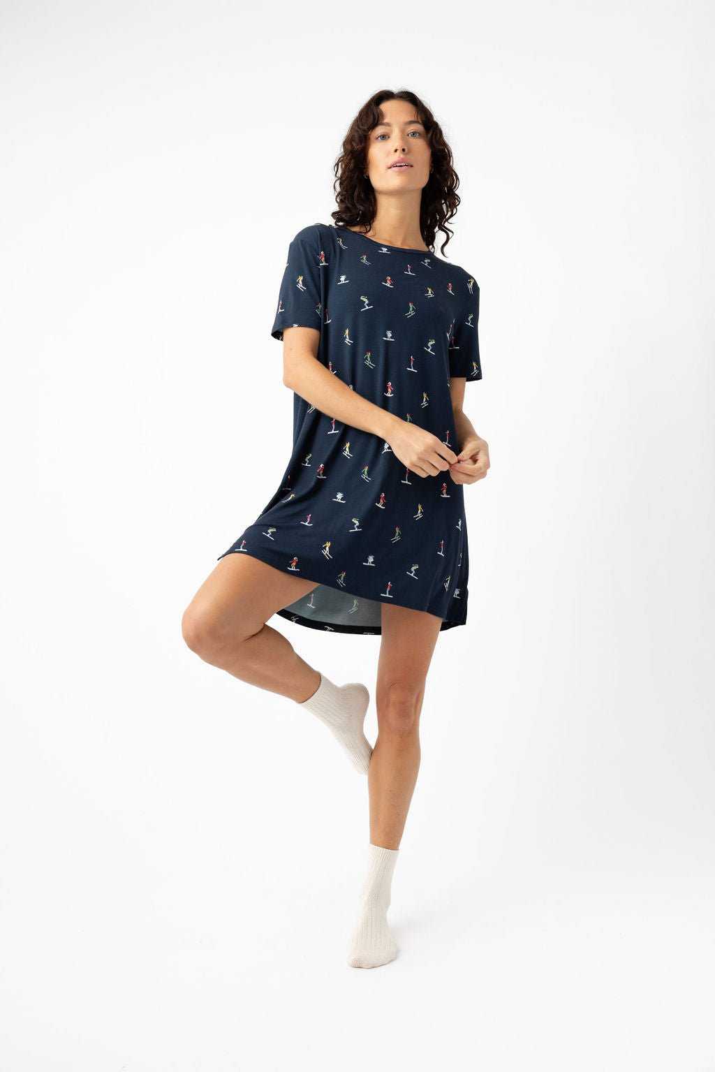 A woman stands on one leg against a white background, wearing the Cozy Earth Women's Bamboo Stretch-Knit Sleep Dress with small colorful prints and white socks. She looks slightly upward, holding the edge of her dress with one hand. |Color:Skiers Navy