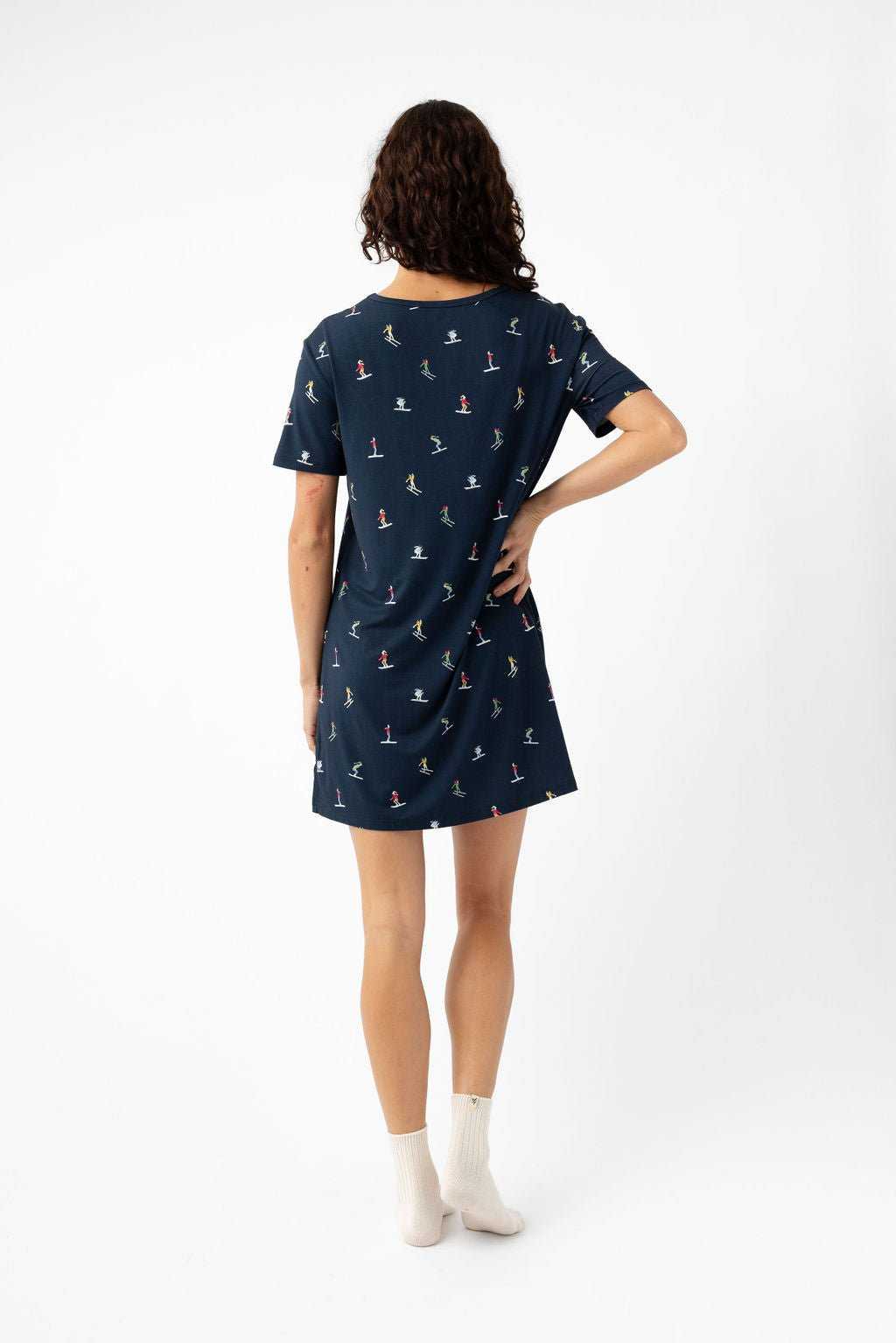 A woman with curly hair stands with her back to the camera, wearing the Cozy Earth Women's Bamboo Stretch-Knit Sleep Dress in navy blue with a small colorful print and white ankle socks, against a plain white background. |Color:Skiers Navy