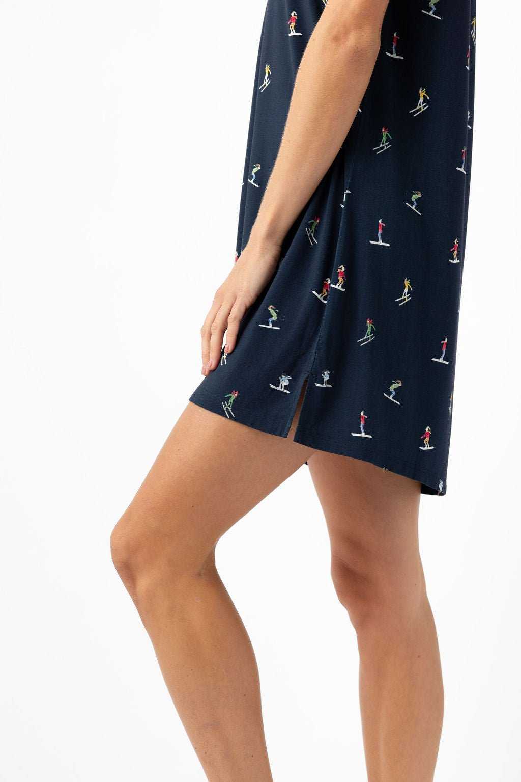 A person wearing the Cozy Earth Women's Bamboo Stretch-Knit Sleep Dress in navy blue with small, colorful skier prints stands against a white background. The dress has a small slit near the hem and only the lower half of their body is visible. |Color:Skiers Navy