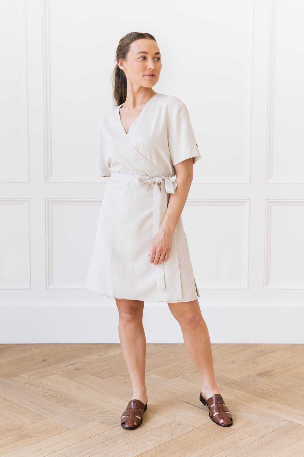 A woman stands in a white room with wooden flooring. She is wearing the Cozy Earth Women's Coastal Comfort Wrap Dress in Sand, paired with brown sandals. She gazes off to the side with her hands relaxed at her sides. |Color:Sand