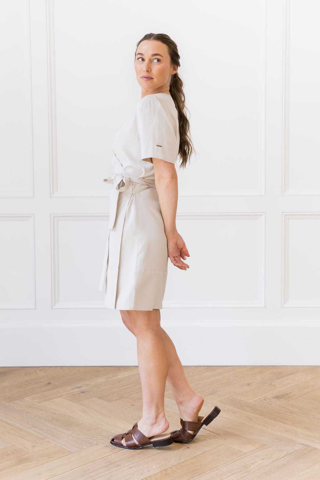 A woman wearing the Women's Cozy Earth Coastal Comfort Wrap Dress from Cozy Earth and brown sandals stands with her body turned slightly to the side. She has long brown hair tied back and holds her hands behind her. The background features a white paneled wall and light wooden flooring. |Color:Sand