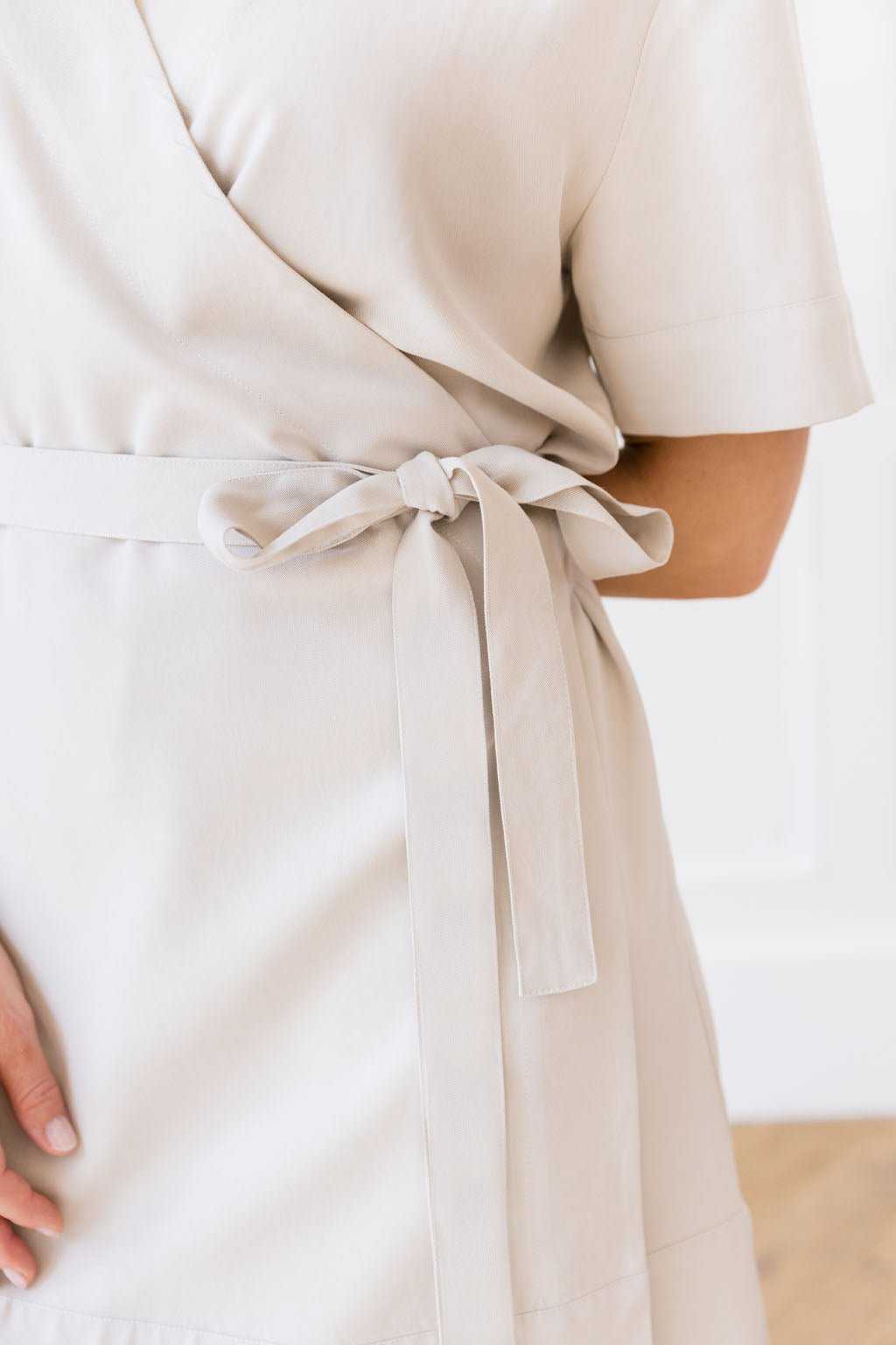 Close-up of a person wearing the Women's Cozy Earth Coastal Comfort Wrap Dress tied with a bow at the waist. The individual, captured standing sideways, rests their left hand gently on the back of their hip. The minimal and bright background highlights the fine details of this Cozy Earth dress. |Color:Sand