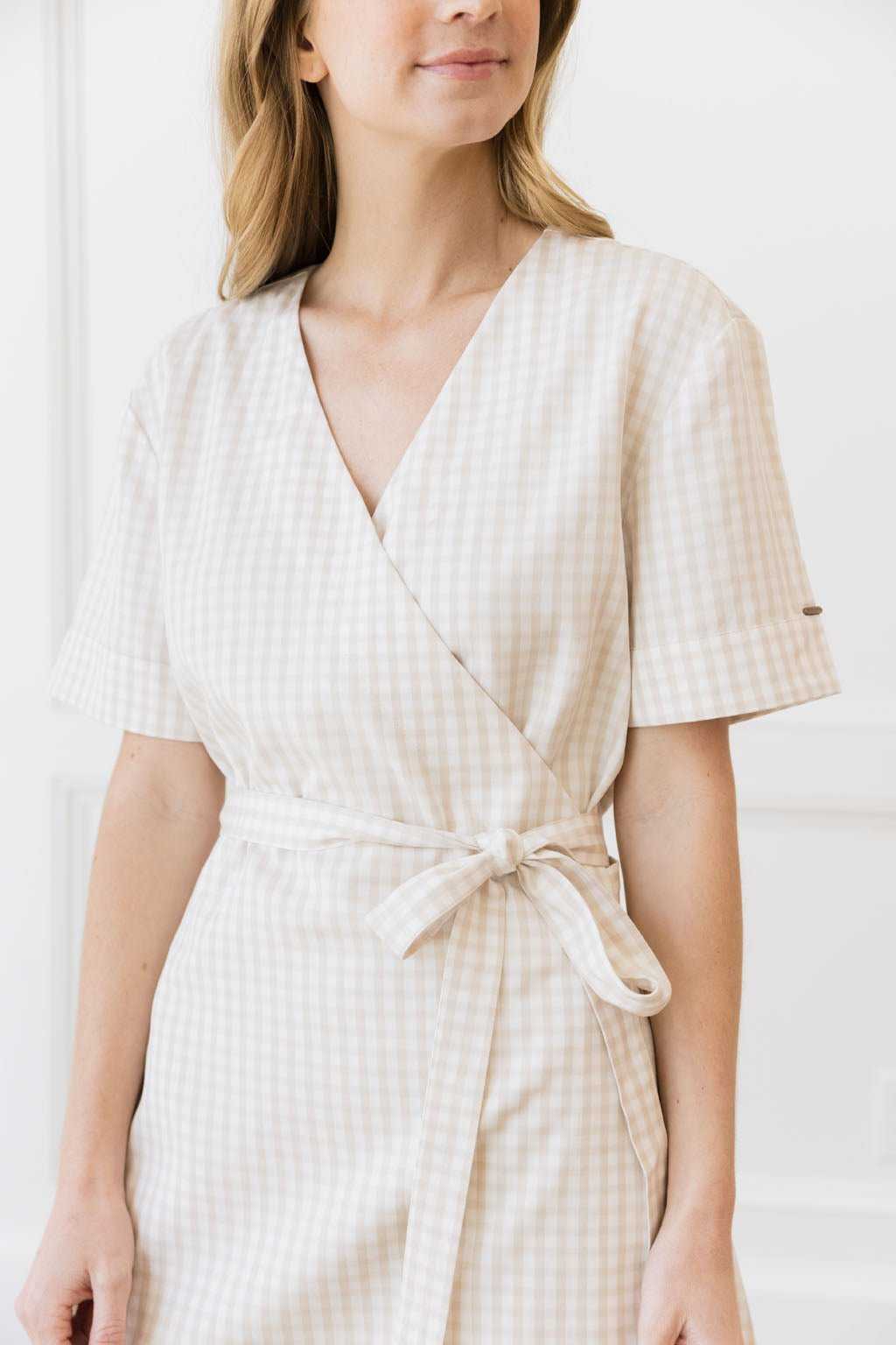 A woman in a beige and white checkered Cozy Earth Coastal Comfort Wrap Dress from Cozy Earth stands against a white background. The dress features short sleeves and a waist tie. Only the upper part of her body is visible, with her face partially out of the frame. |Color:Sunkissed Gingham