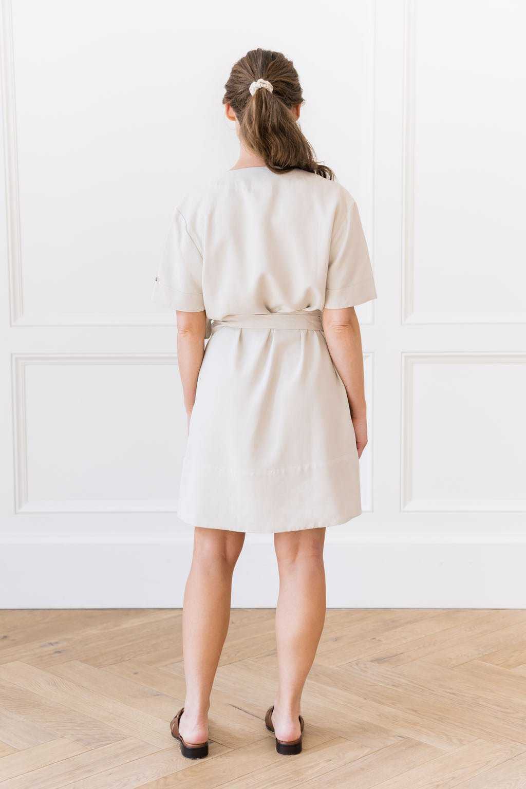 A woman stands with her back to the camera, wearing the Women's Cozy Earth Coastal Comfort Wrap Dress by Cozy Earth, which is short-sleeved and belted at the waist in a light beige color. Her dark hair is tied back in a ponytail with a white scrunchie. The background features a white wall with decorative paneling and a light wood floor. |Color:Sand