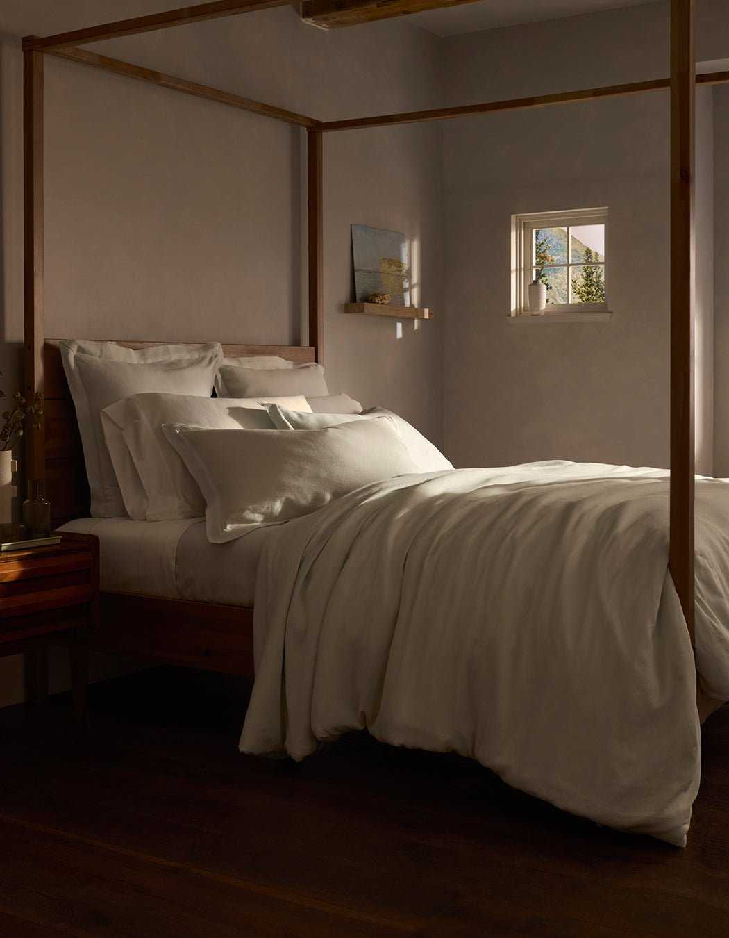A cozy bedroom features a wooden four-poster bed with layered white bedding, including the Linen Duvet Cover by Cozy Earth. Warm light from a small window softly illuminates the neutral tones, creating an inviting atmosphere.|Color:Mist