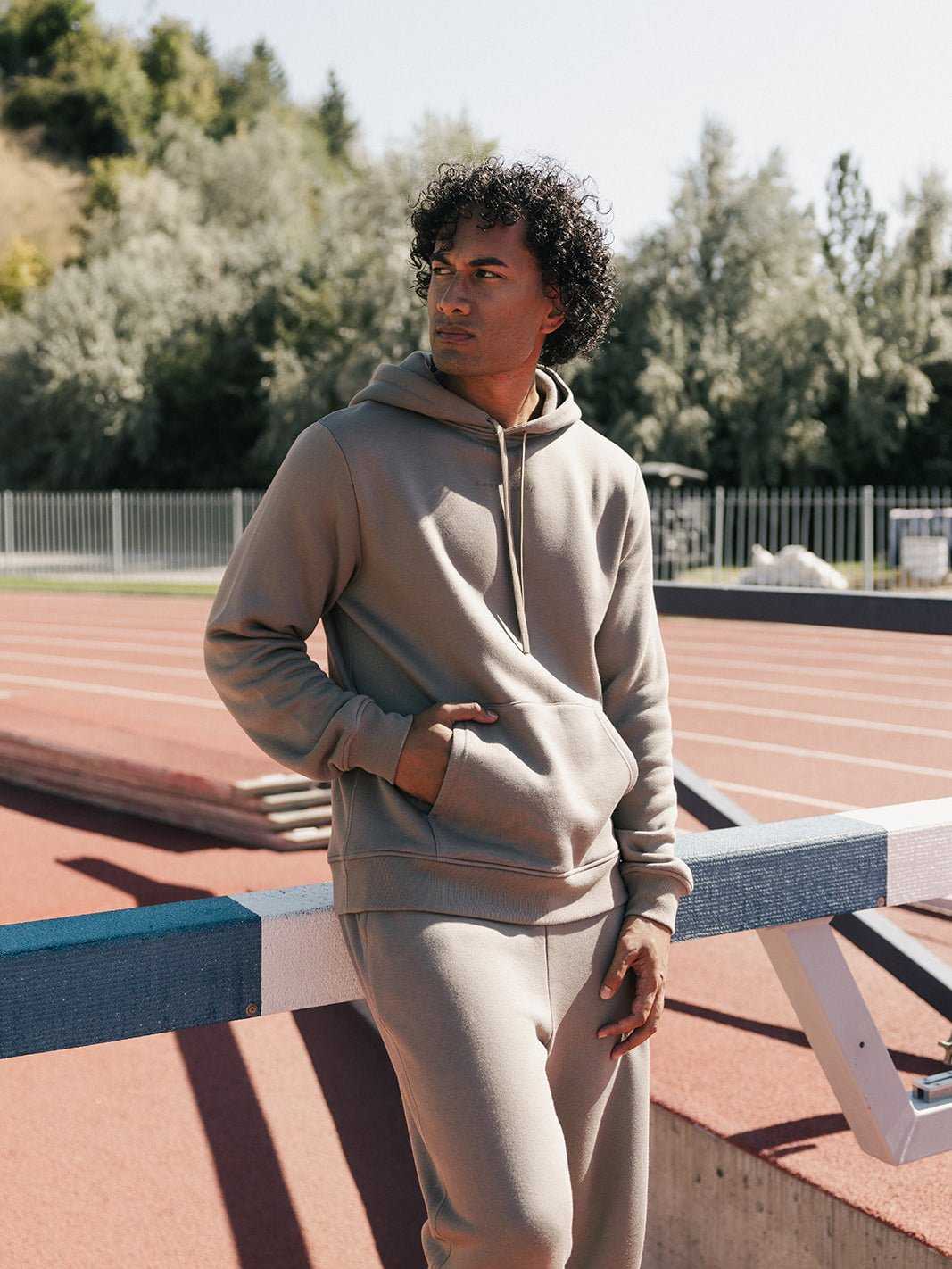 A person with curly hair wearing a beige Men's CityScape Hoodie and matching sweatpants from Cozy Earth stands near a hurdle on an outdoor track field. They have one hand in their pocket and gaze thoughtfully into the distance, with trees and a fence visible in the background. |Color:Ebony