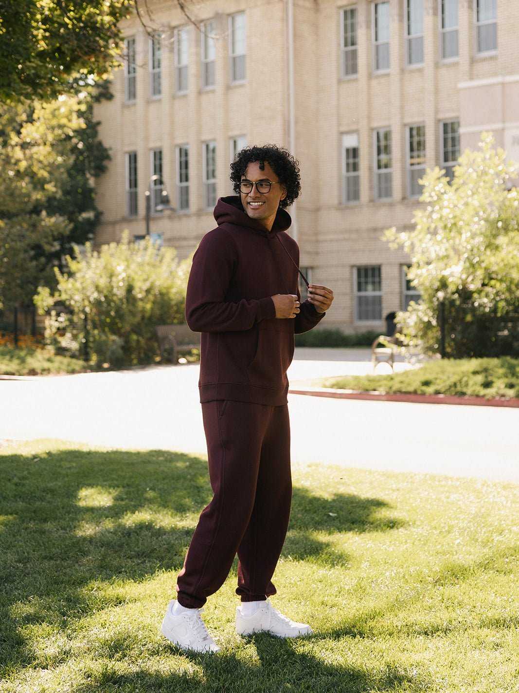 A person with curly hair, wearing glasses, and dressed in a maroon Men's CityScape Hoodie and matching sweatpants from Cozy Earth, stands smiling on a green lawn in front of a beige brick building. Bright sunlight casts shadows, and trees and bushes are visible in the background. |Color:Burgundy