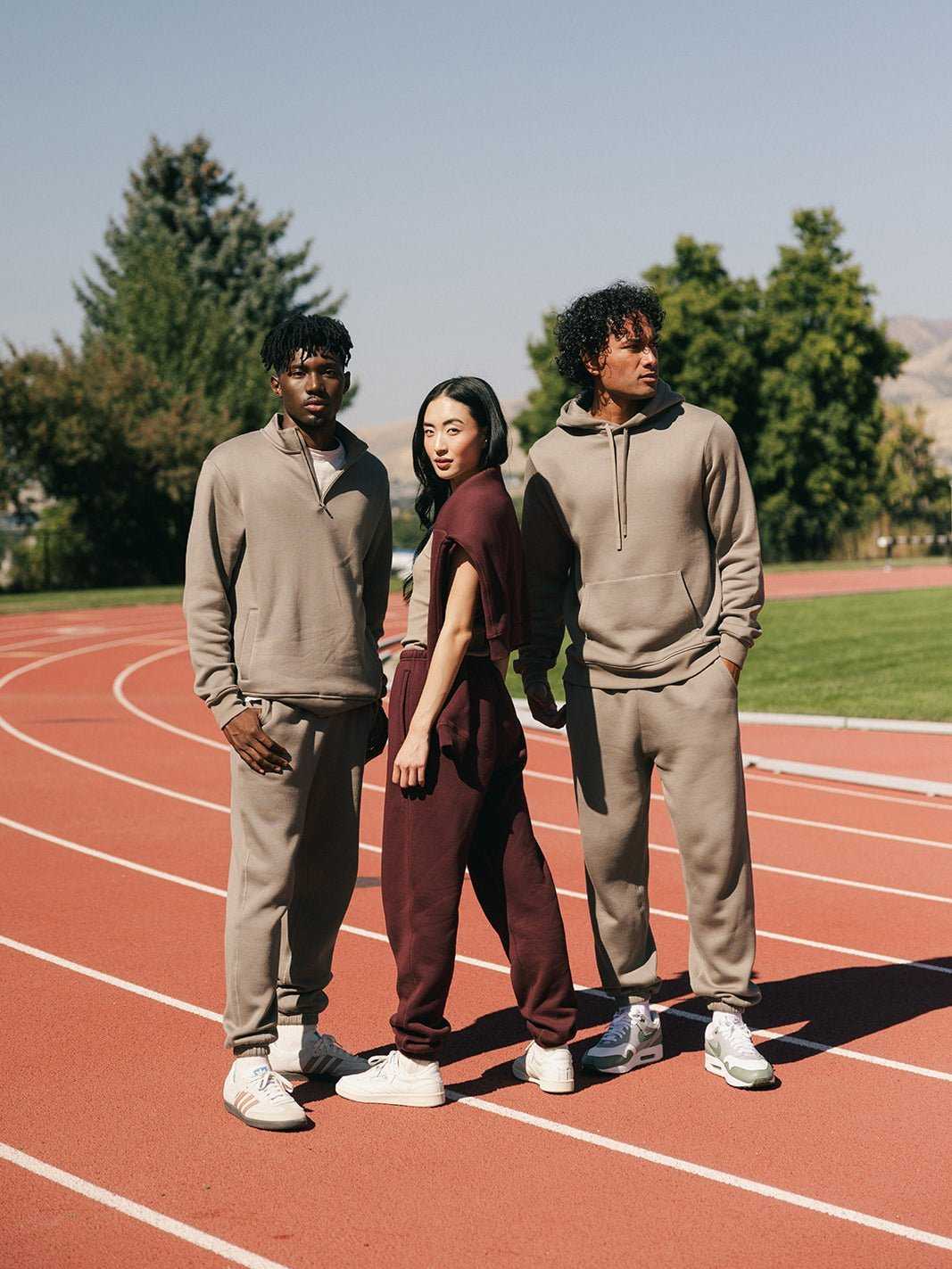 Three people stand on an outdoor track under a clear sky. Two individuals wear beige Men's CityScape Sweatpants from Cozy Earth, while the person in the middle wears maroon Men's CityScape Sweatpants from Cozy Earth. They all wear white sneakers and pose confidently with trees and a distant hill in the background. |Color:Ebony