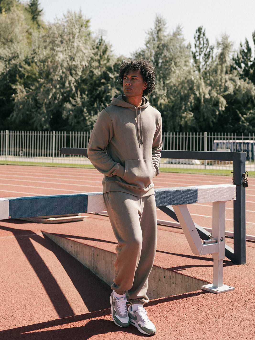 A person wearing a Cozy Earth Men's CityScape Sweatpant and a matching tan hoodie leans against a hurdle on an outdoor track, gazing away from the camera. Trees and a metal fence are visible in the background beneath a clear sky. |Color:Ebony