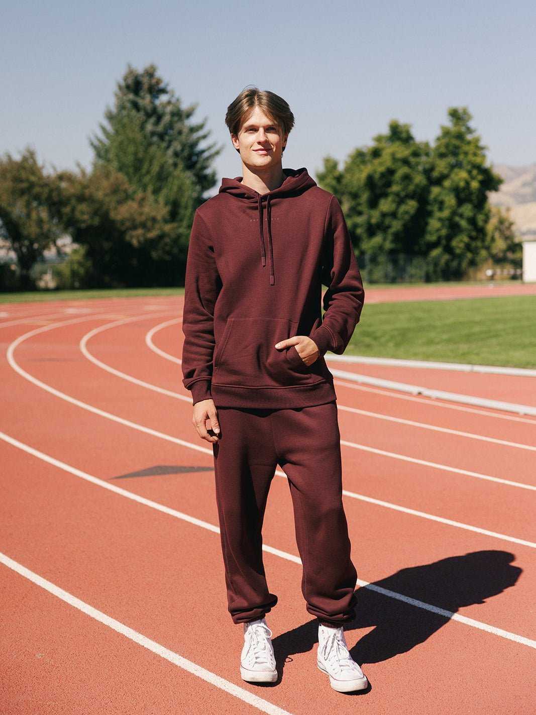 A young man stands on an outdoor running track on a sunny day, dressed in Cozy Earth's Men's CityScape Sweatpants and matching burgundy hoodie, paired with white sneakers. Green trees and a clear sky can be seen in the background. He is smiling and facing the camera. |Color:Burgundy