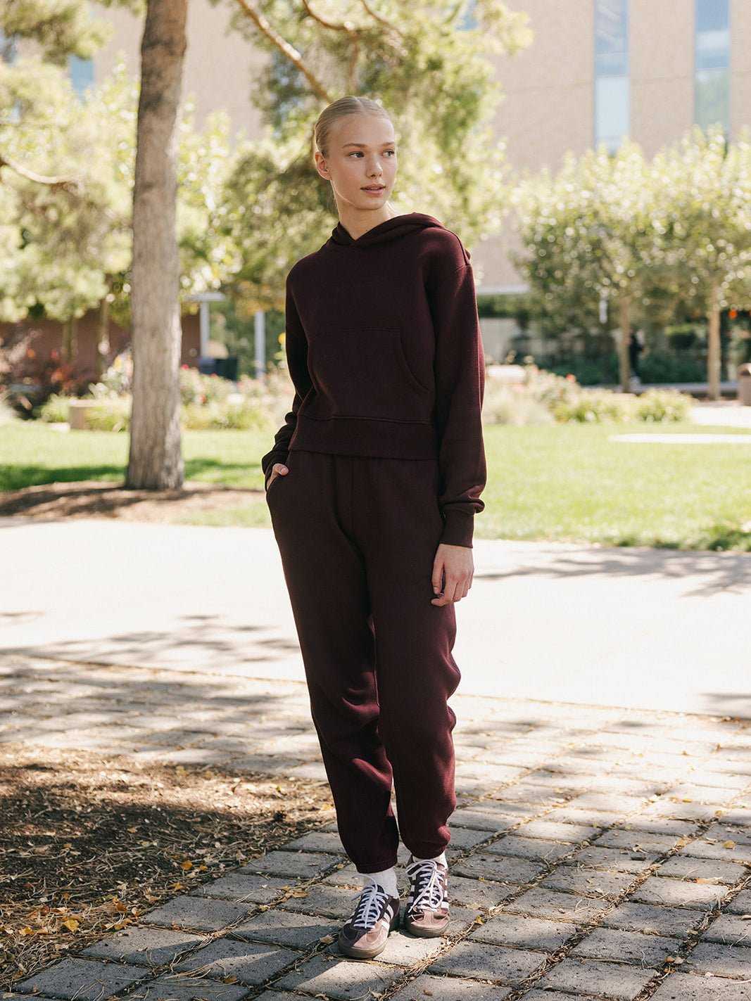 A person with light hair is standing outdoors on a paved pathway surrounded by trees and greenery. They are wearing a matching dark burgundy set, including the Women's CityScape Cropped Hoodie from Cozy Earth, and looking to the side with a relaxed expression. The background includes a building and sunlight filtering through the leaves. |Color:Burgundy