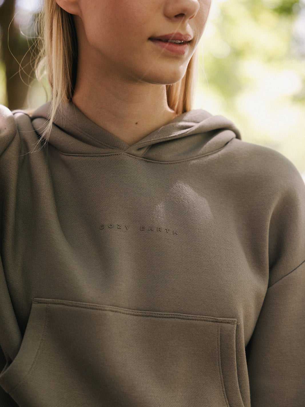 A person wearing a Women's CityScape Cropped Hoodie in olive-green, embellished with the subtle text "Cozy Earth," stands in soft, natural light. The background is blurred, featuring greenery that suggests an outdoor setting. The photo is cropped at the shoulders, with only part of the person's face visible. |Color:Ebony