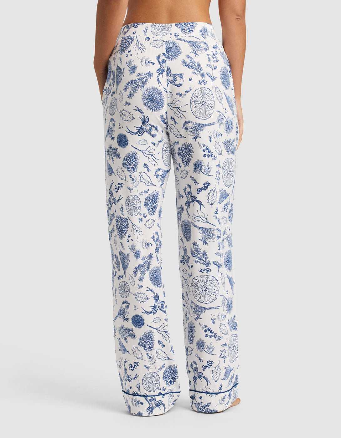 A person stands with their back to the camera wearing Cozy Earth Women's Bamboo Stretch-Knit Pajama Pant in white with a blue floral print. The pants are loose-fitting with pockets, showing only the lower back and legs against a plain background.|Color:Winter Toile Pacific