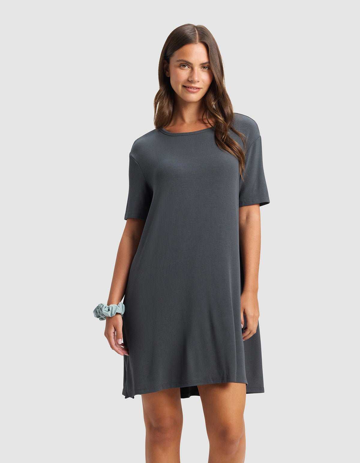 A woman with long brown hair wears the Cozy Earth Women’s Bamboo Rib-Knit Boyfriend Sleep Dress in dark gray and a light blue scrunchie on her wrist. She smiles softly against a plain light gray background.|Color:Graphite