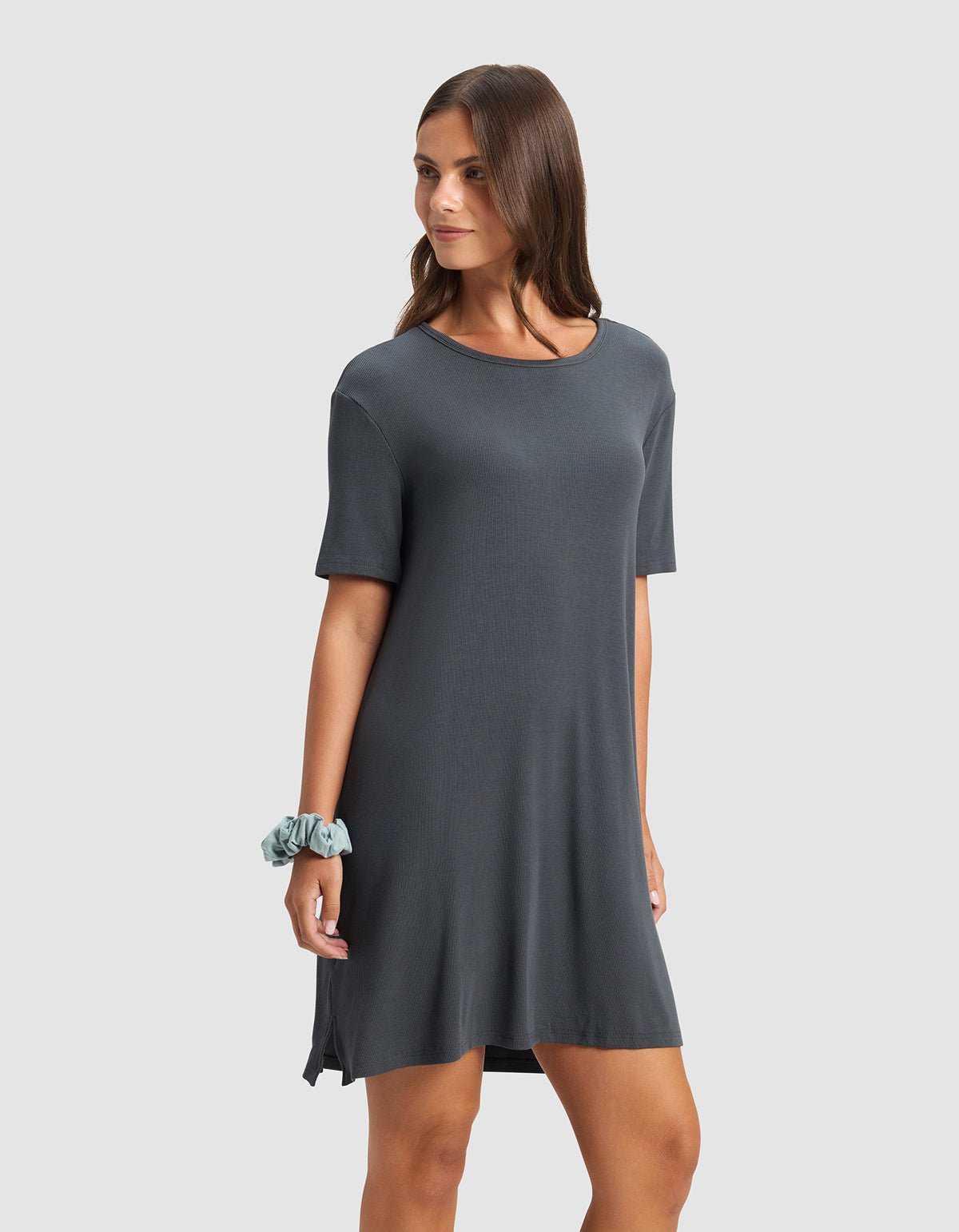 A woman wears the Cozy Earth Women’s Bamboo Rib-Knit Boyfriend Sleep Dress in dark gray, her long brown hair down and a light blue scrunchie on her left wrist, standing against a plain light gray background.|Color:Graphite