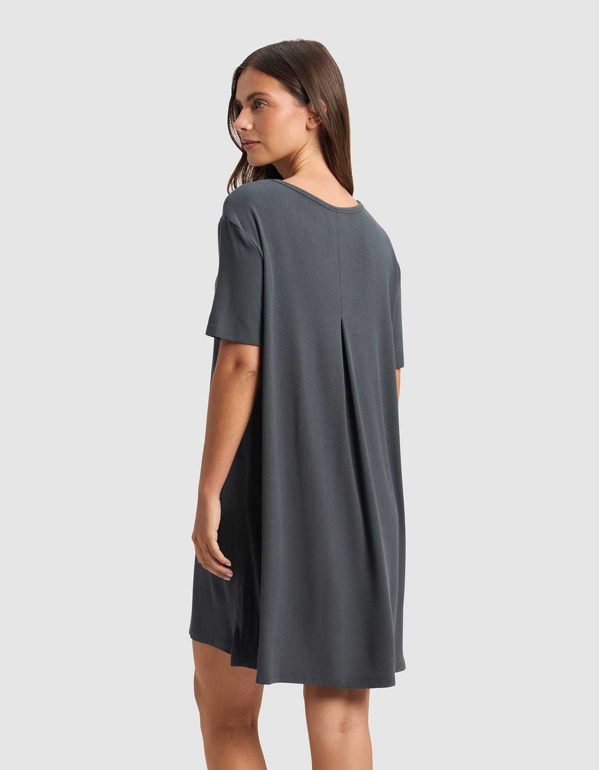 A woman with straight brown hair models the Cozy Earth Women’s Bamboo Rib-Knit Boyfriend Sleep Dress in charcoal gray, viewed from the back. The loose-fitting, short-sleeve dress features a center pleat and falls above the knees against a plain light gray background.|Color:Graphite