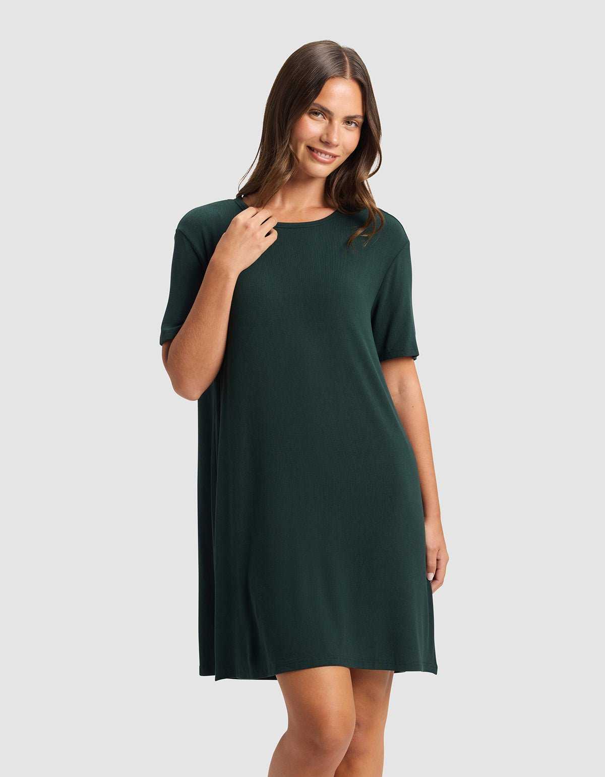 A woman with long brown hair smiles and poses in the Cozy Earth Women’s Bamboo Rib-Knit Boyfriend Sleep Dress, a dark green, short-sleeve, knee-length style, against a plain light gray background.|Color:Spruce