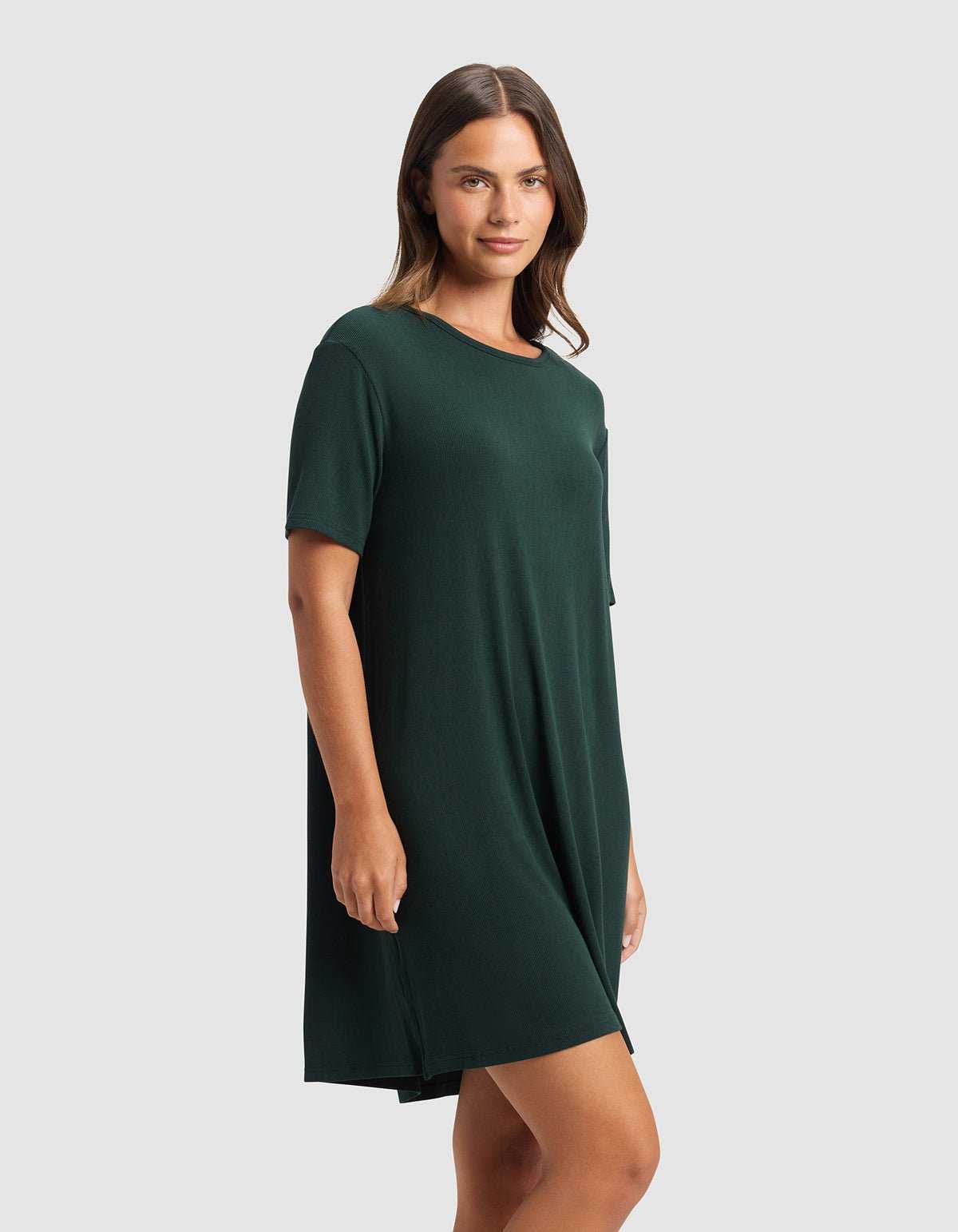 A woman with straight brown hair wears the Cozy Earth Women’s Bamboo Rib-Knit Boyfriend Sleep Dress in dark green, standing against a plain light gray background and looking at the camera with a slight smile.|Color:Spruce