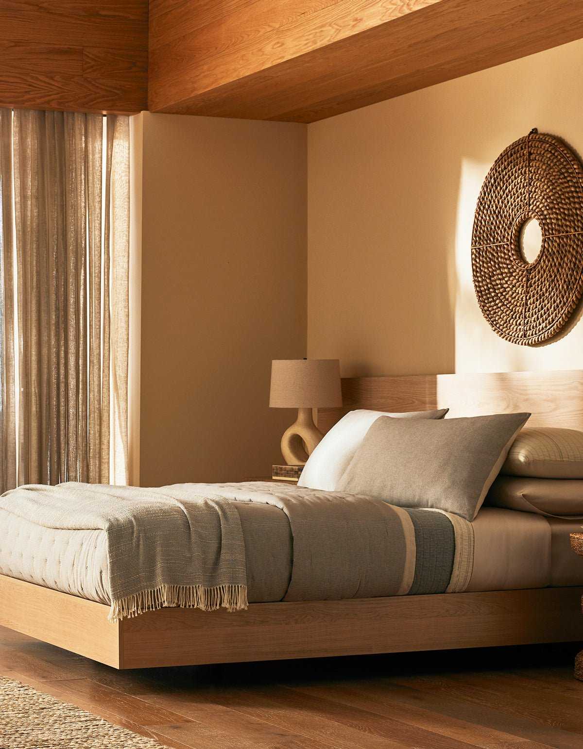 A modern bedroom features Cozy Earth's Baja Quilt Bundle on a wooden platform bed, with neutral tones, a woven wall hanging, a beige lamp on a nightstand, and sunlight filtering through sheer curtains onto wood floors.|Color:Coal Stripe/Striped Alabaster