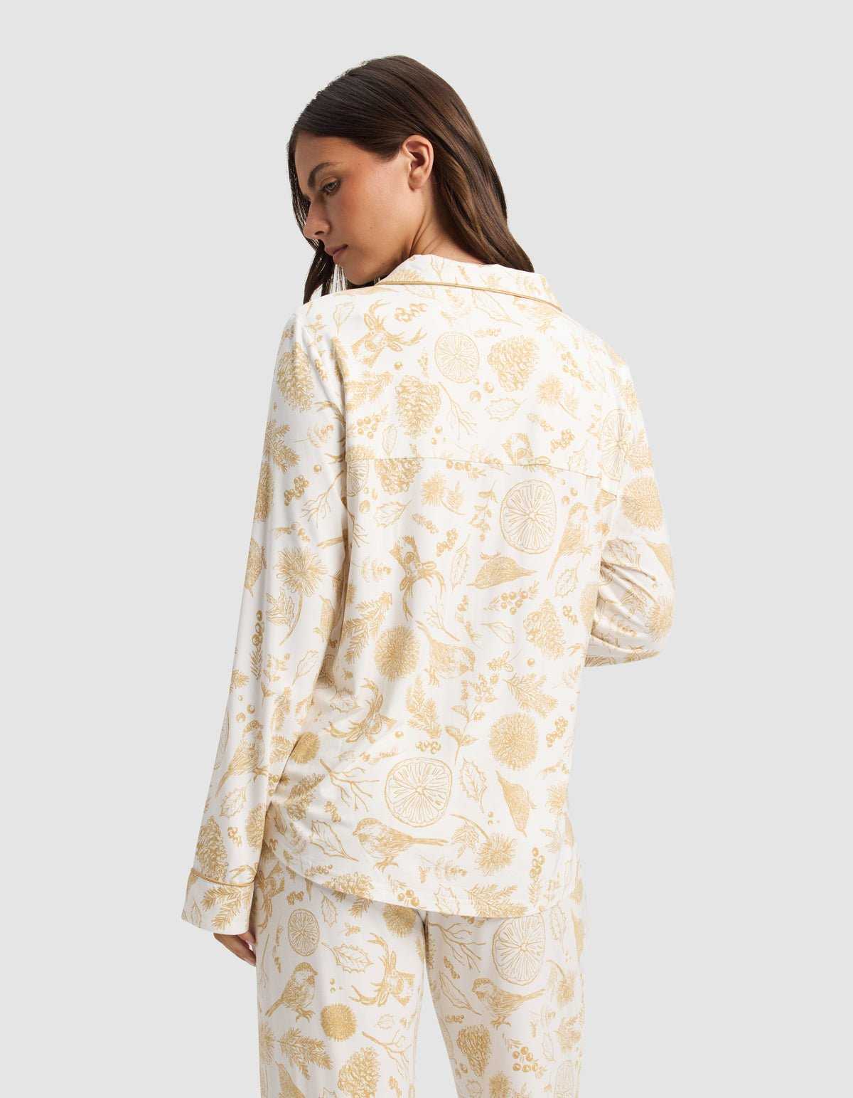 A woman with long brown hair is seen from behind wearing the Cozy Earth Women's Bamboo Stretch-Knit Long Sleeve Pajama Top in white with a yellow botanical and fruit print, standing against a light gray background.|Color:Winter Toile Dijon