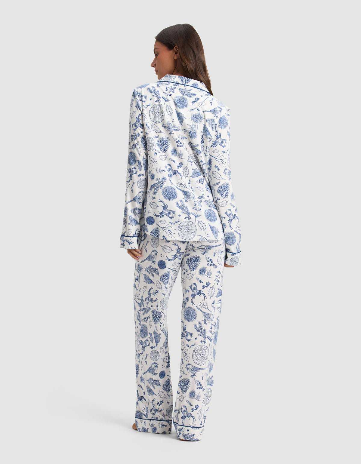A woman stands barefoot facing away, wearing the Cozy Earth Women's Bamboo Stretch-Knit Long Sleeve Pajama Set in a blue and white botanical print against a plain light gray background.|Color:Winter Toile Pacific