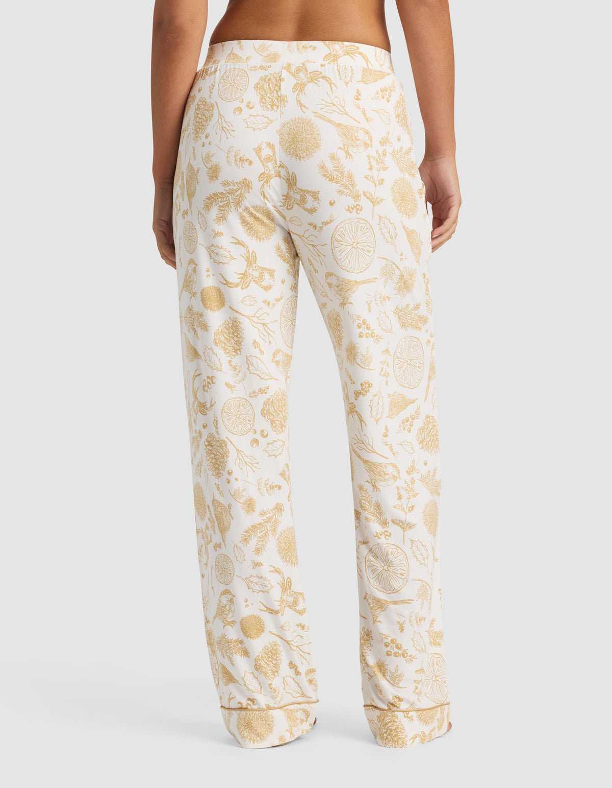 A person models the back view of the Cozy Earth Women's Bamboo Stretch-Knit Pajama Pant, featuring a gold botanical and citrus fruit print on white, shown in a relaxed fit against a plain background.|Color:Winter Toile Dijon