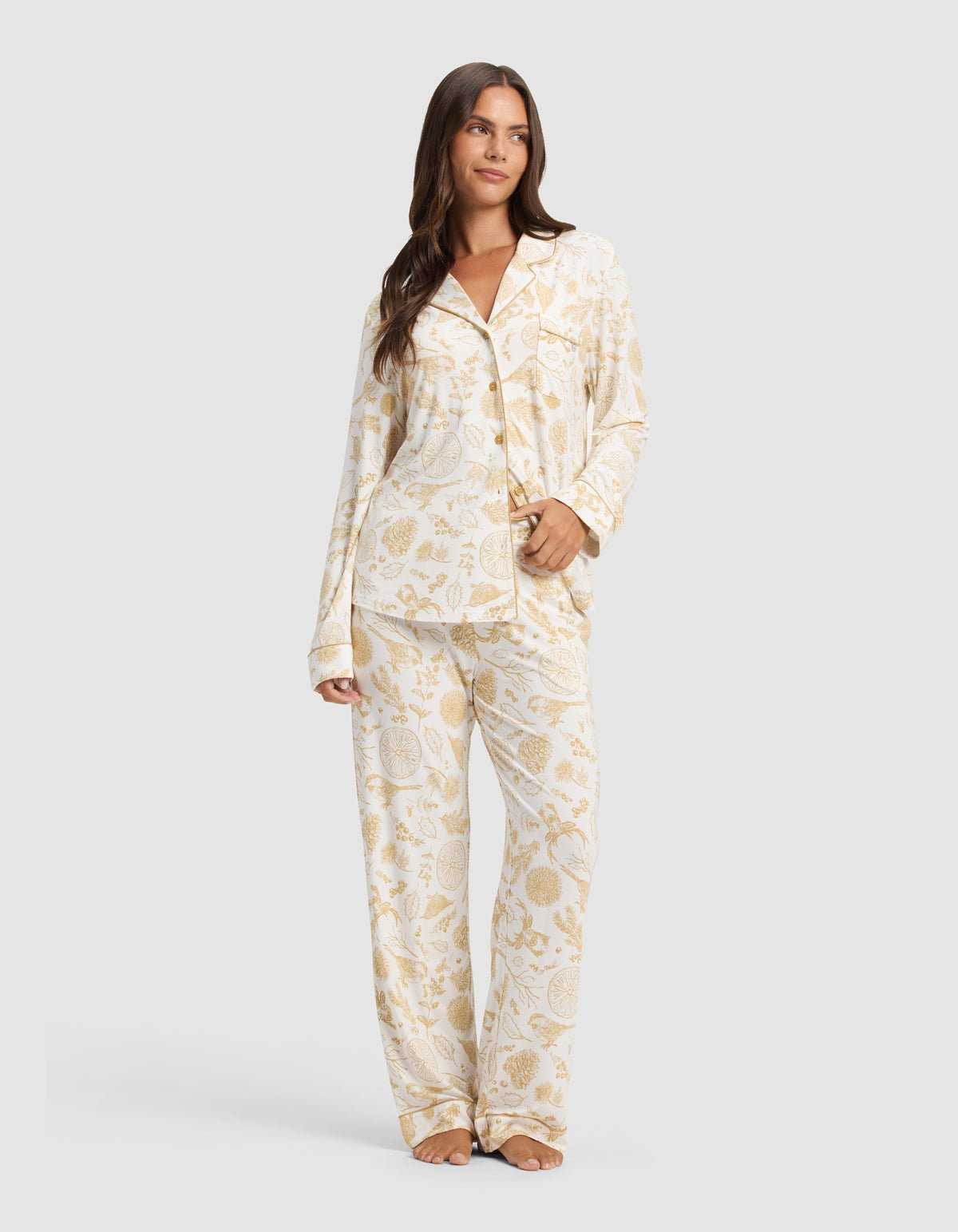 A woman stands barefoot against a light gray background wearing Cozy Earth's Women's Bamboo Stretch-Knit Pajama Pant in cream with a gold botanical print, paired with a matching long-sleeve button-up shirt.|Color:Winter Toile Dijon