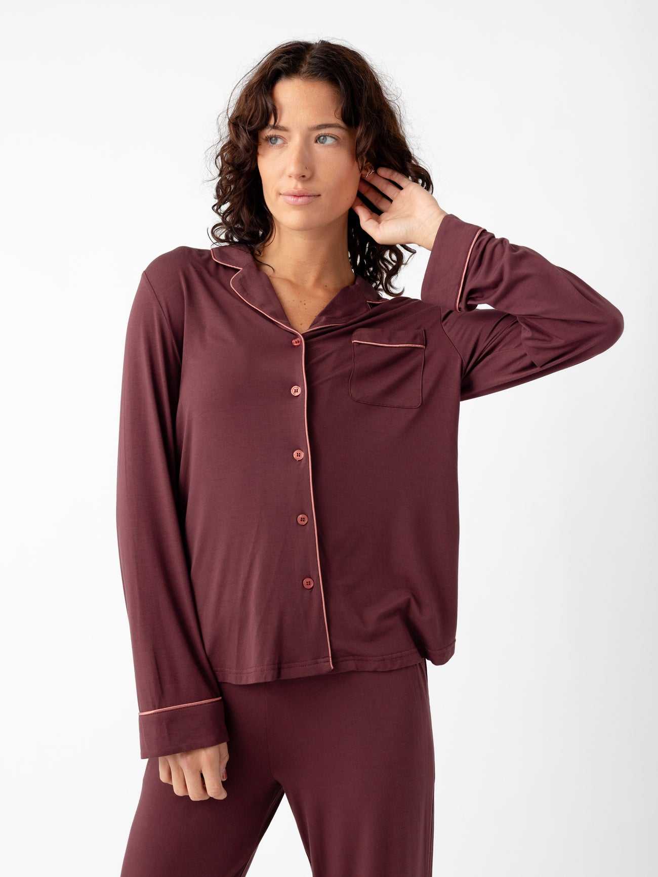 Wearing the Last Chance Women's Bamboo Stretch-Knit Long Sleeve Pajama Set by Cozy Earth, a person with maroon pajamas featuring a button-down front and chest pocket touches their hair against a plain white background. |Color:Burgundy