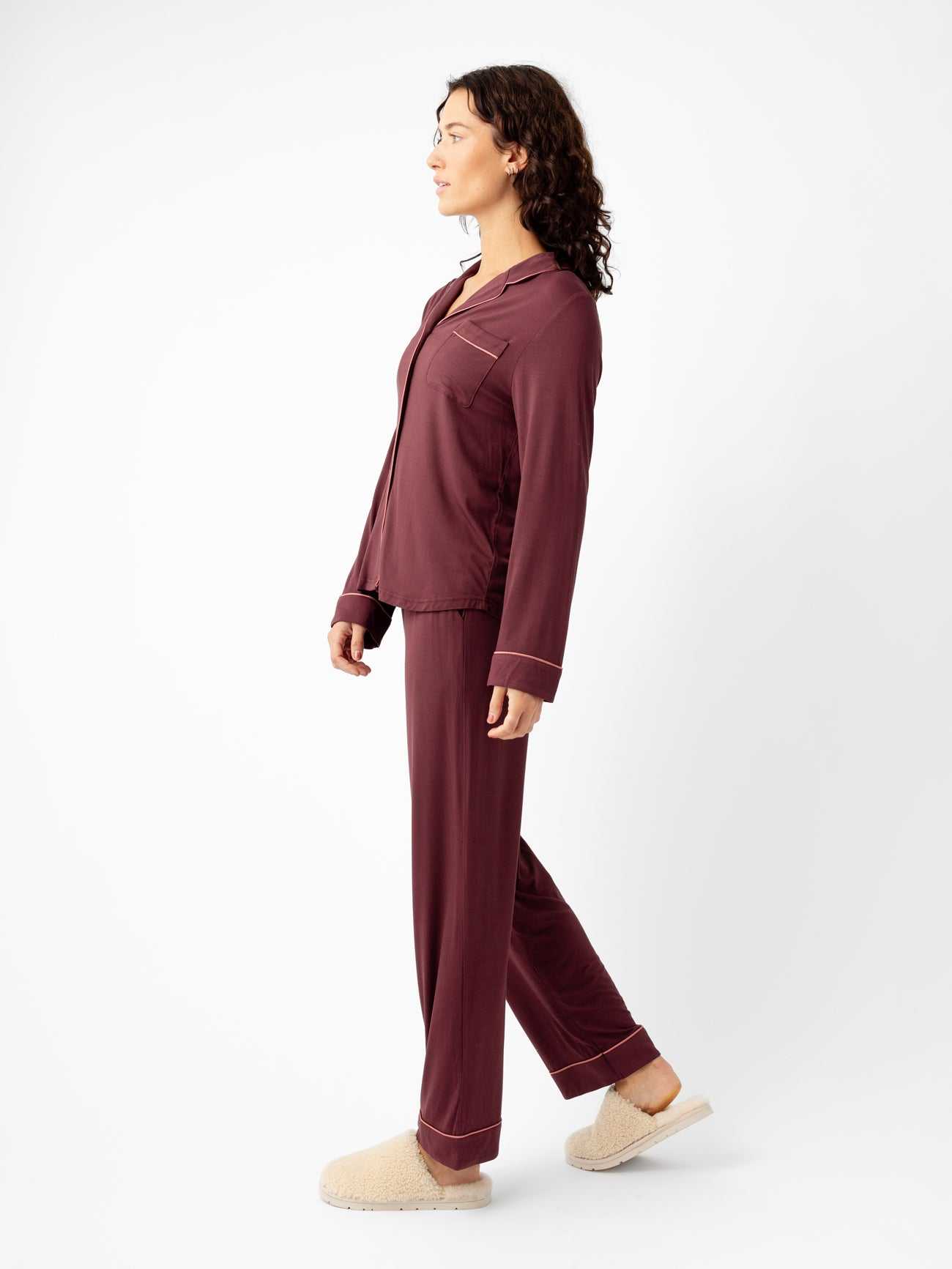 A person with wavy hair stands in profile against a plain white background, wearing the Last Chance Women's Bamboo Stretch-Knit Long Sleeve Pajama Set by Cozy Earth in maroon and beige slippers. |Color:Burgundy