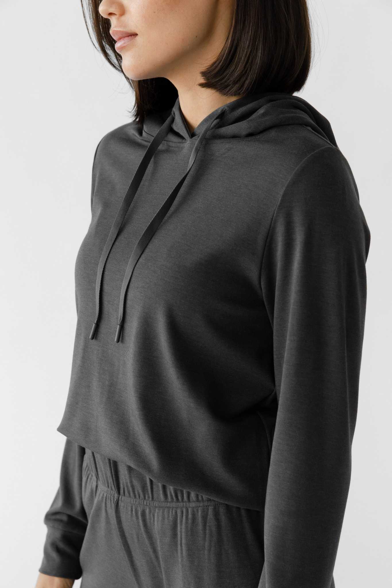 A woman wearing a grey Cozy Earth Brushed Bamboo Hoodie with a drawstring and sleeve cuffs. The hoodie has an embroidered logo on the front. |Color:Charcoal