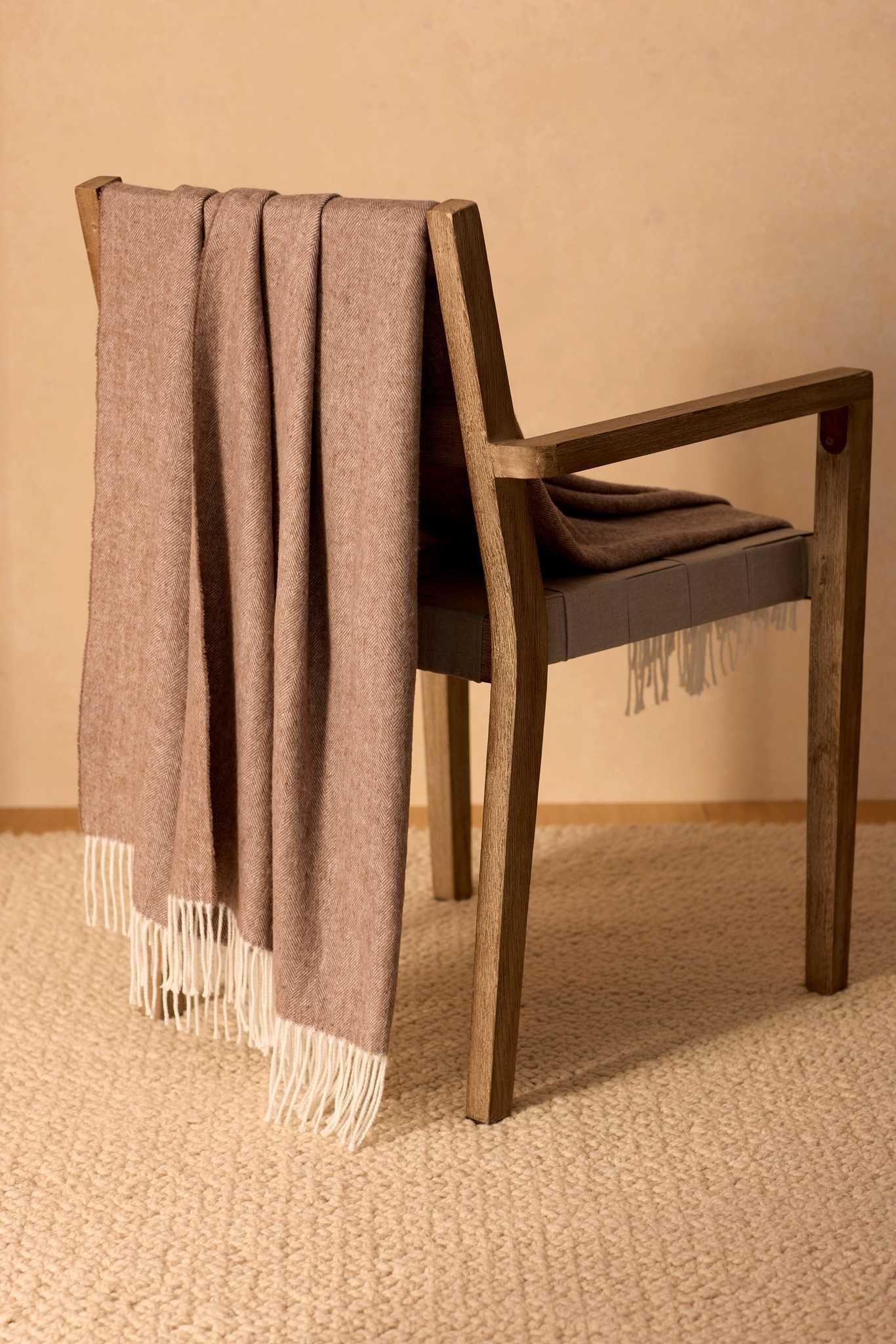 A wooden chair with the Cozy Earth Bamboo Herringbone Tassel Throw in taupe, featuring white fringed ends, is draped over the backrest and sits on a textured beige carpet against a light tan wall.|Color:Truffle