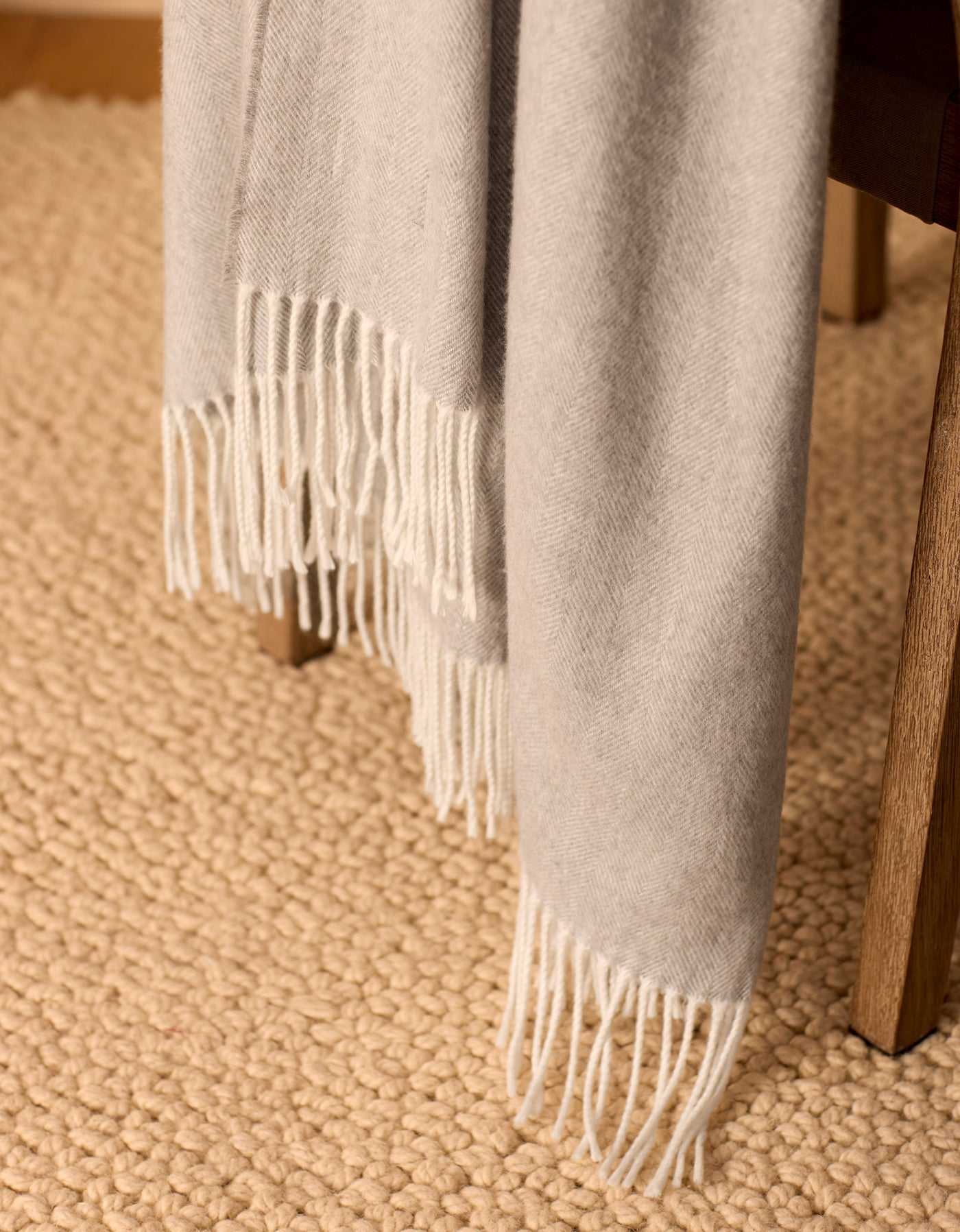 The Bamboo Herringbone Tassel Throw by Cozy Earth, in light gray, is draped over a wooden chair with a textured beige rug placed on the floor beneath.|Color:Cloud