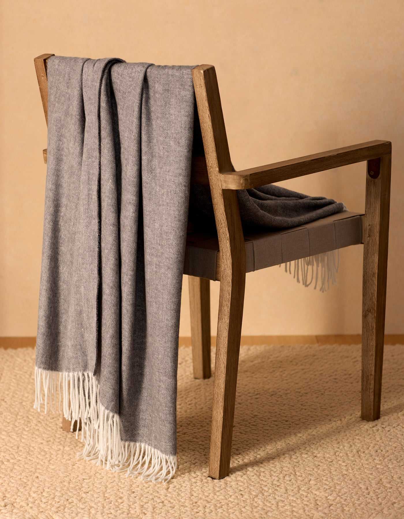 The Bamboo Herringbone Tassel Throw by Cozy Earth, in soft gray with white fringe, is draped over a wooden chair on a beige textured rug against a light tan wall. |Color:Shadow