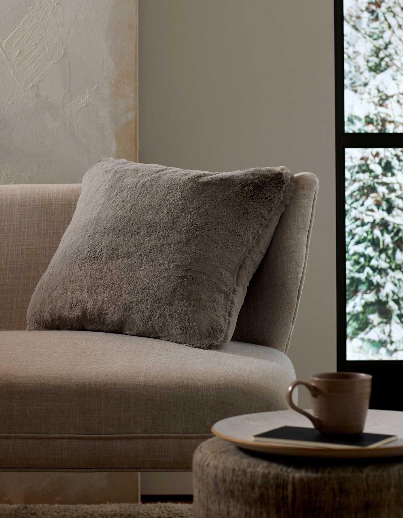A cozy beige couch with a large, soft Cuddle Pillow by Cozy Earth rests beside a round table holding a ceramic mug and coaster. Sunlight pours through the window, revealing snowy trees outside.|Color:Dove Grey