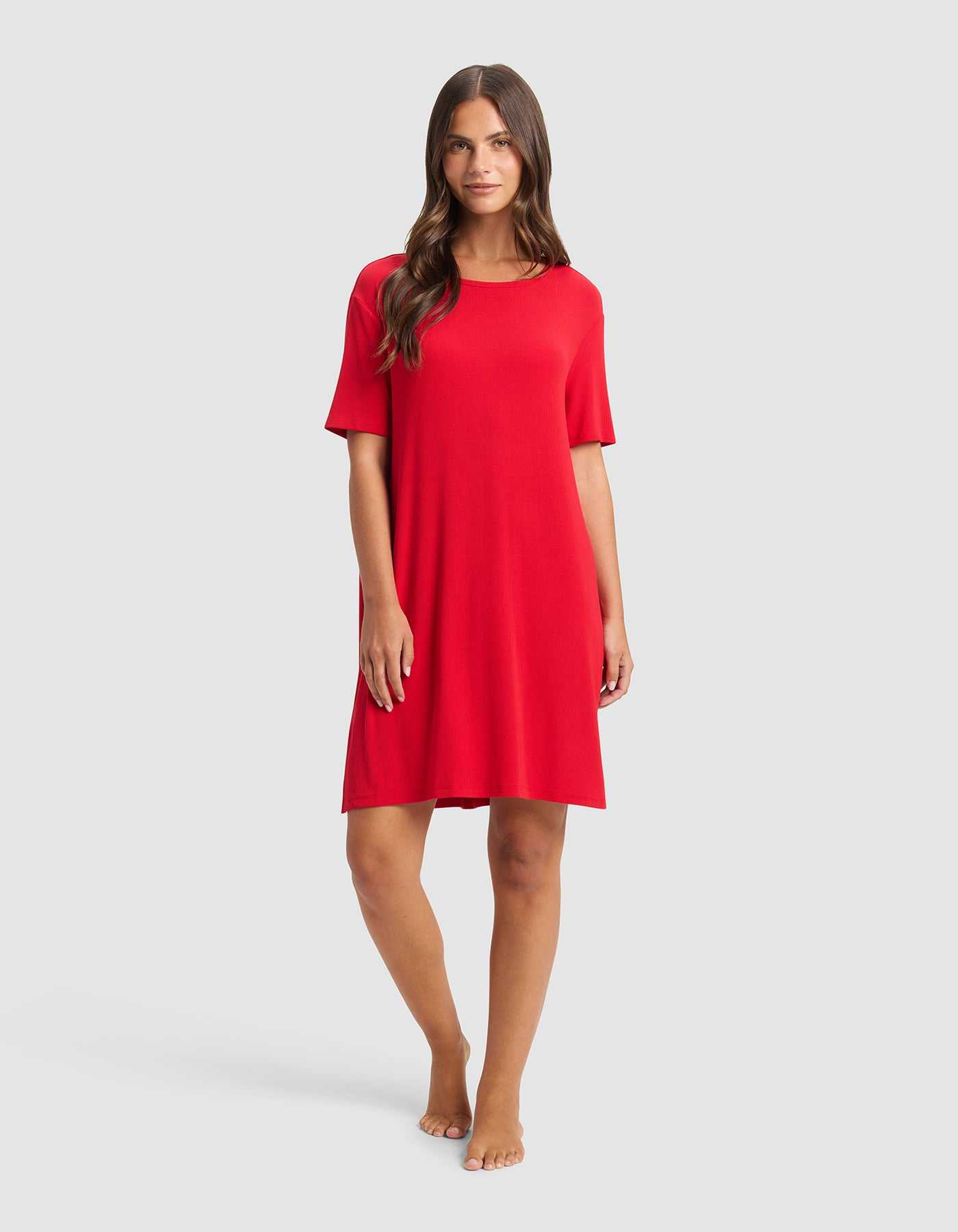 A woman with long brown hair stands barefoot against a light gray background, wearing the Cozy Earth Women’s Bamboo Rib-Knit Boyfriend Sleep Dress in red, featuring short sleeves and a loose fit that falls just above her knees.|Color:Crimson