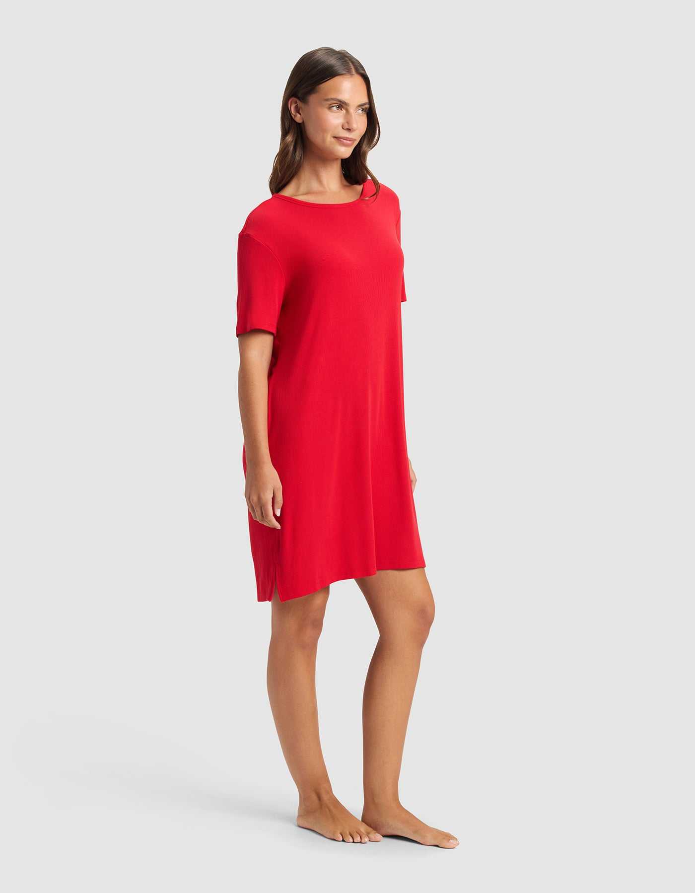 A woman stands barefoot against a light gray background, wearing the Cozy Earth Women’s Bamboo Rib-Knit Boyfriend Sleep Dress in red. She smiles softly and looks to the side.|Color:Crimson