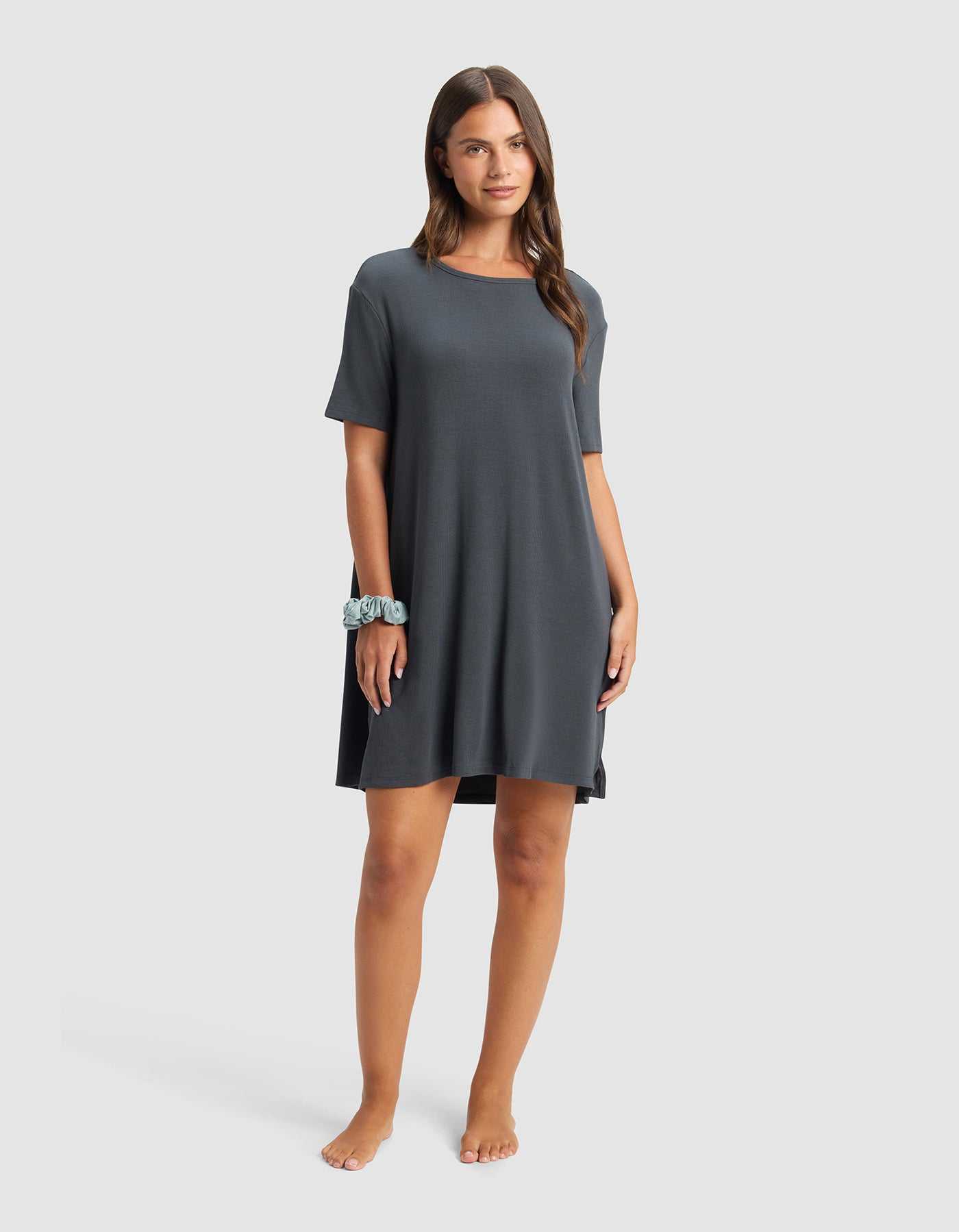 A woman with long brown hair stands barefoot against a plain background, wearing the Cozy Earth Women’s Bamboo Rib-Knit Boyfriend Sleep Dress in dark gray, short-sleeved and loose-fitting, with a blue scrunchie on her wrist.|Color:Graphite