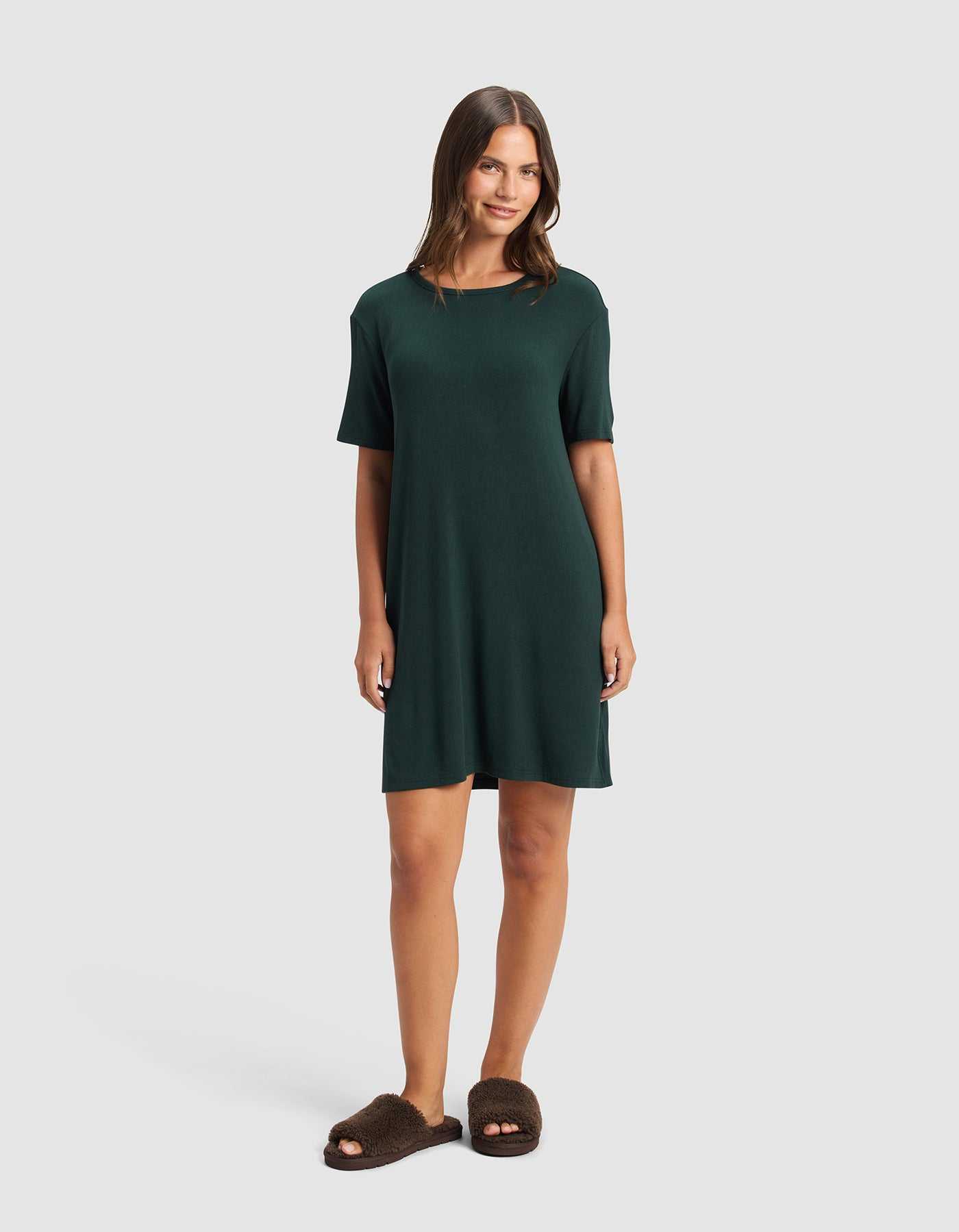 A woman smiles, wearing the Cozy Earth Women's Bamboo Rib-Knit Boyfriend Sleep Dress and brown fuzzy slippers, standing against a plain light gray background.|Color:Spruce