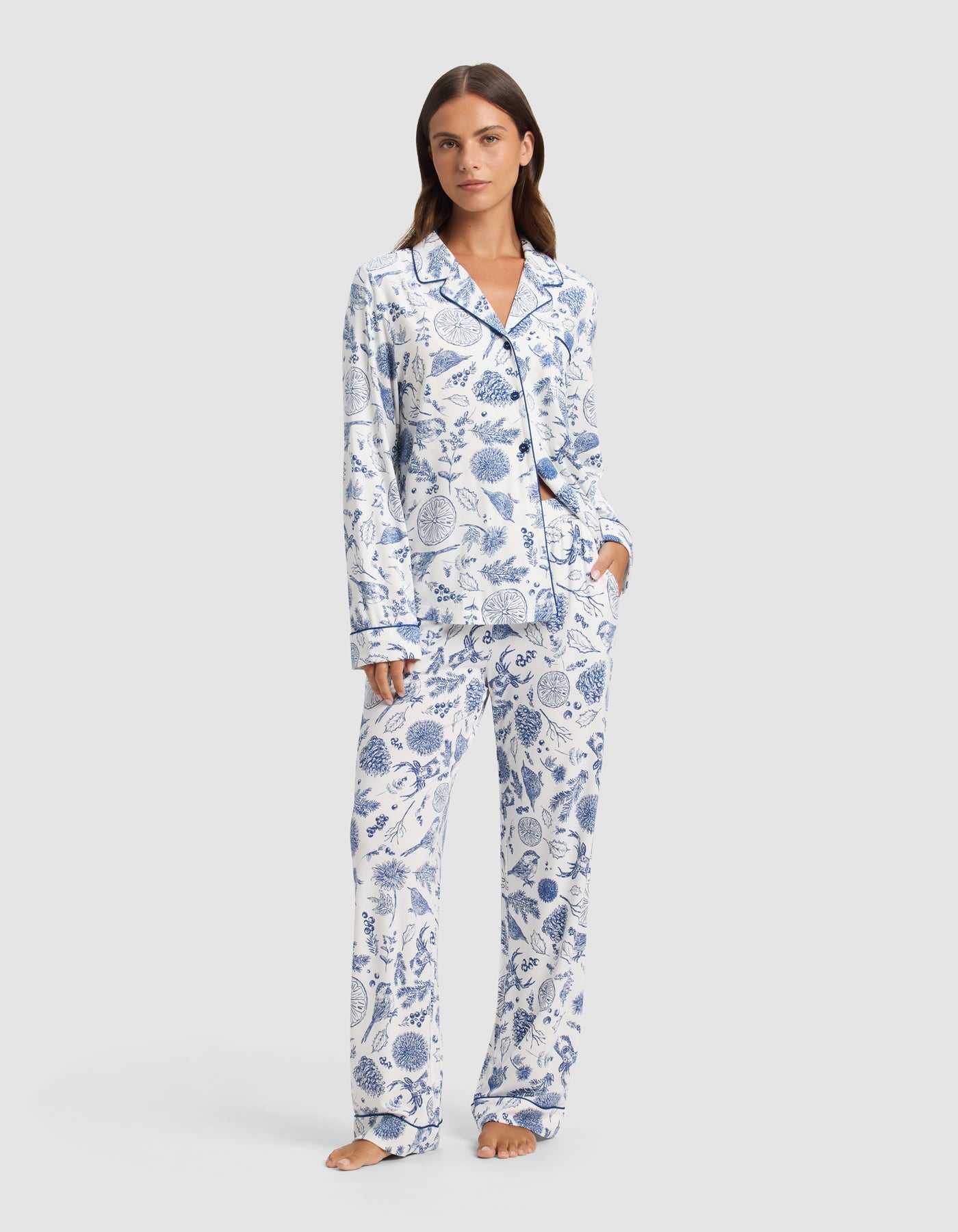 A woman stands barefoot wearing Cozy Earth's Women's Bamboo Stretch-Knit Pajama Pant, featuring a white background with blue botanical print and paired with a matching long-sleeve button-up top.|Color:Winter Toile Pacific