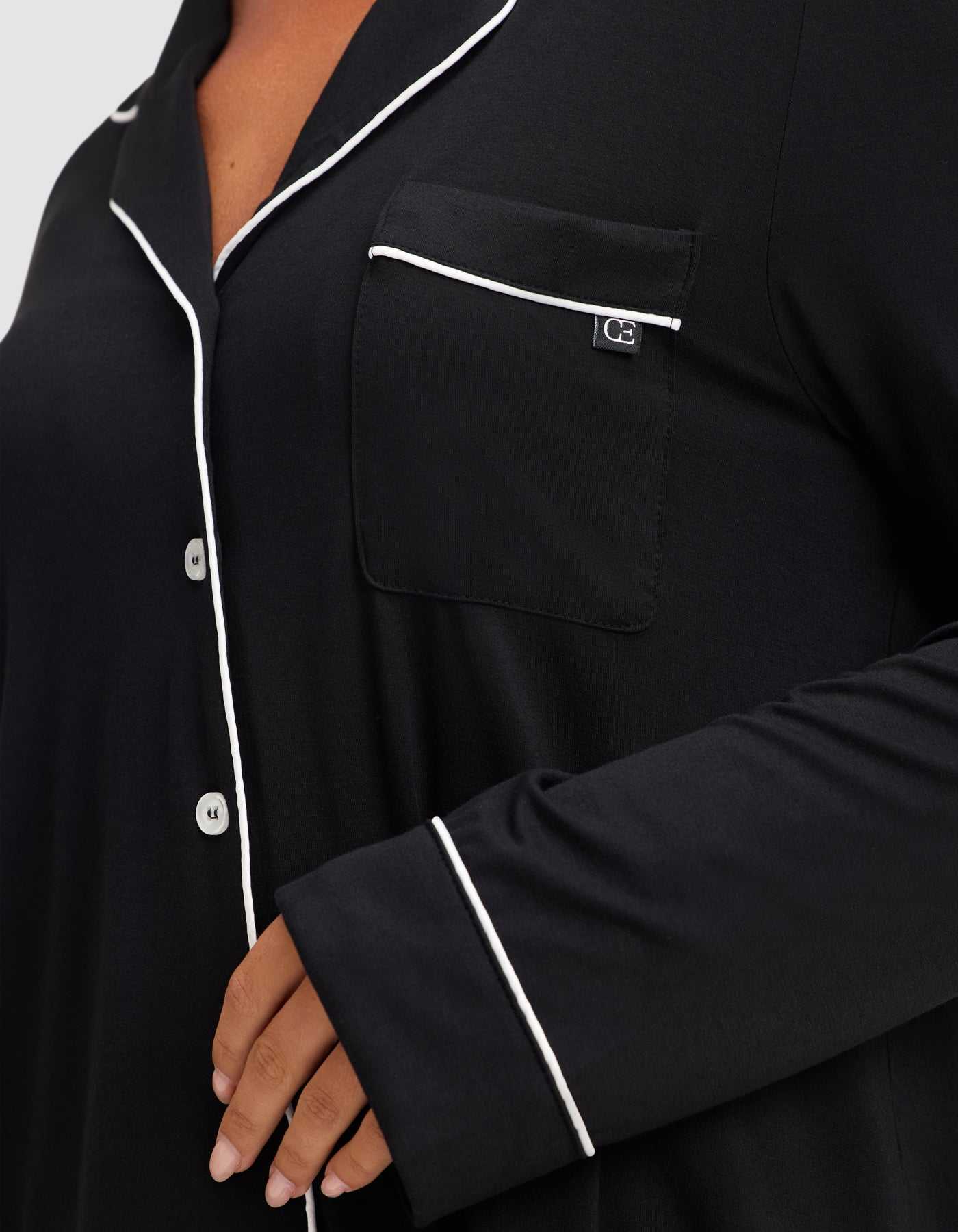 A close-up shows a person wearing the HIDE Women's Bamboo Stretch-Knit Long Sleeve Pajama Top in black, featuring white piping on the collar, pocket, and sleeve, with four white buttons visible and a hand resting near the waist. |Color:Black