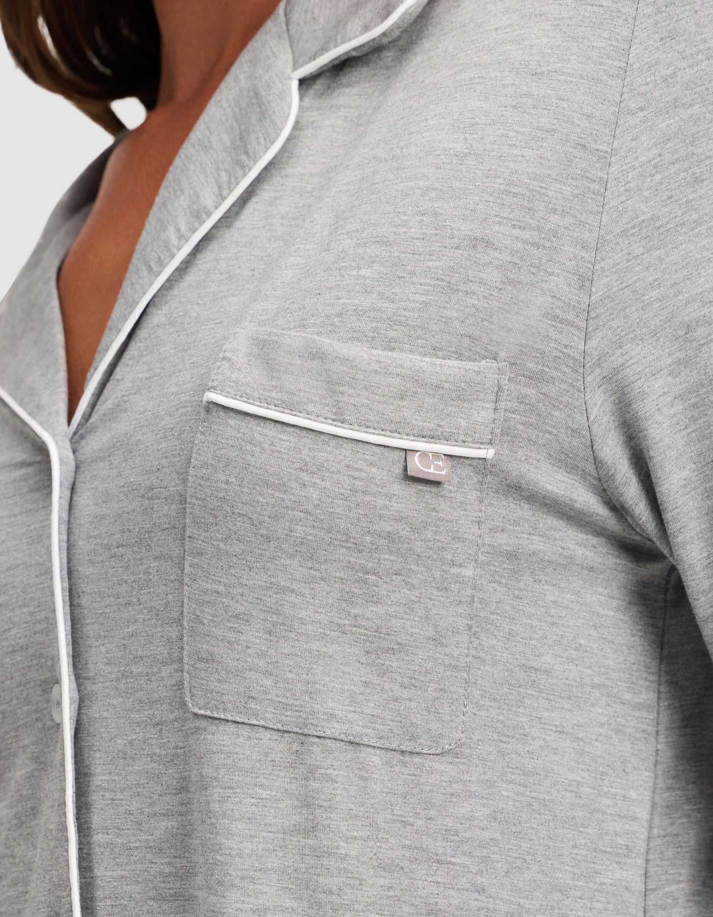 Close-up of a person wearing the HIDE Women's Bamboo Stretch-Knit Long Sleeve Pajama Top in light gray with white piping on the collar, pocket, and trim. The fabric appears soft, and a small tag is visible on the chest pocket's edge. |Color:Grey