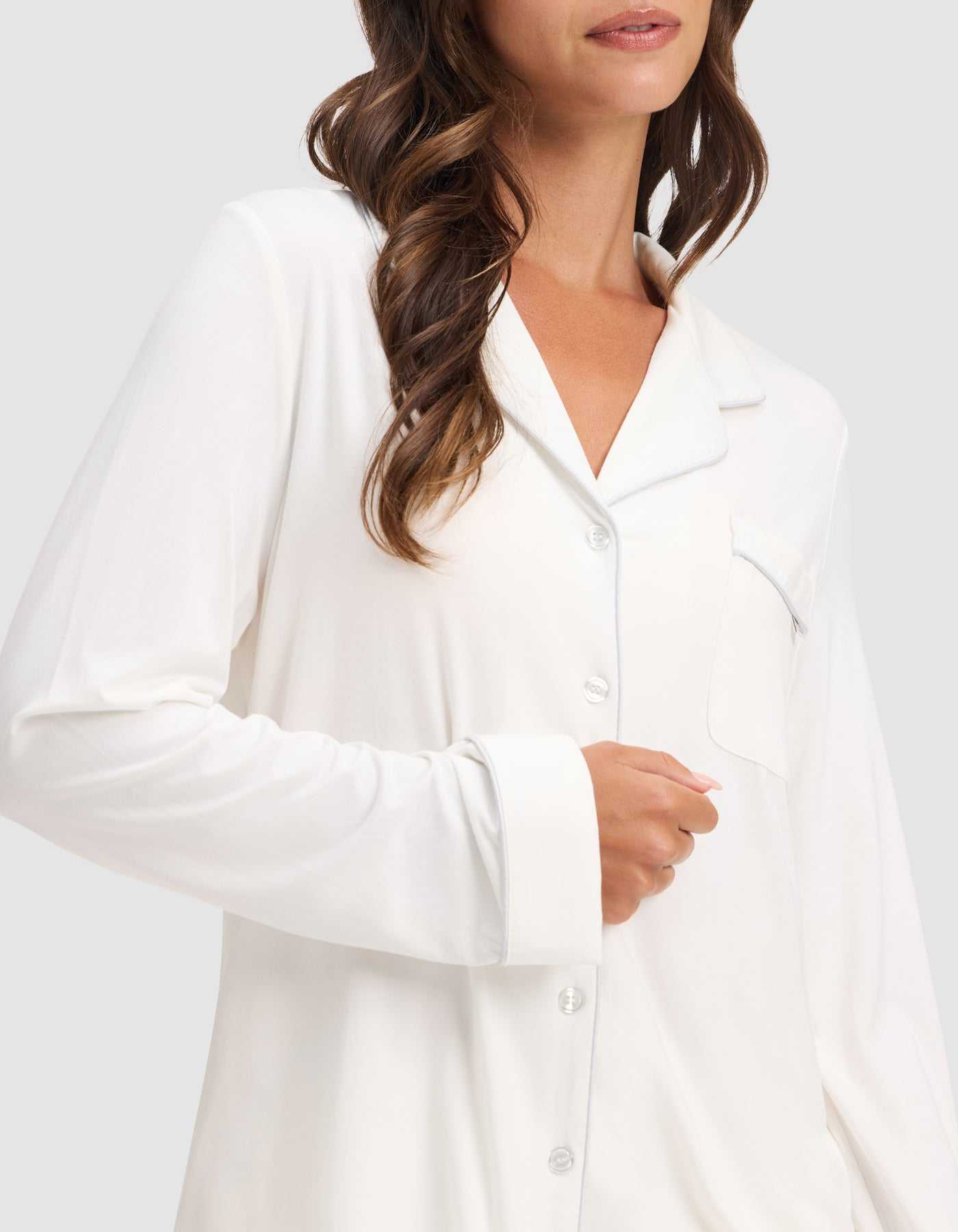 A woman with wavy brown hair wears the HIDE Women's Bamboo Stretch-Knit Long Sleeve Pajama Top, featuring a chest pocket and button-up front. She is shown from shoulders to waist, with her hand resting near the buttons. |Color:Ivory