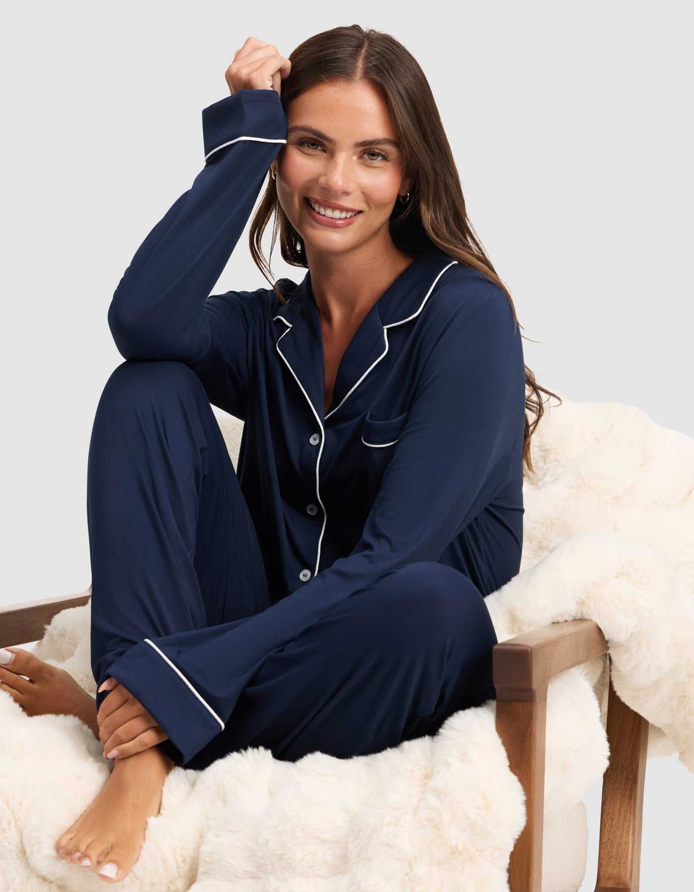 A woman with long brown hair smiles barefoot on a white faux fur chair, wearing the HIDE Women's Bamboo Stretch-Knit Long Sleeve Pajama Top in navy blue with white piping, set against a light gray background. |Color:Navy