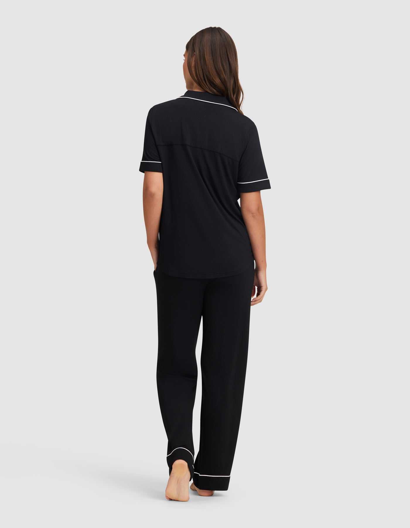 A woman stands barefoot facing away, wearing the Cozy Earth Women's Bamboo Stretch-Knit Short Sleeve & Pant Pajama Set in black with white piping, set against a plain light gray background.|Color:Black