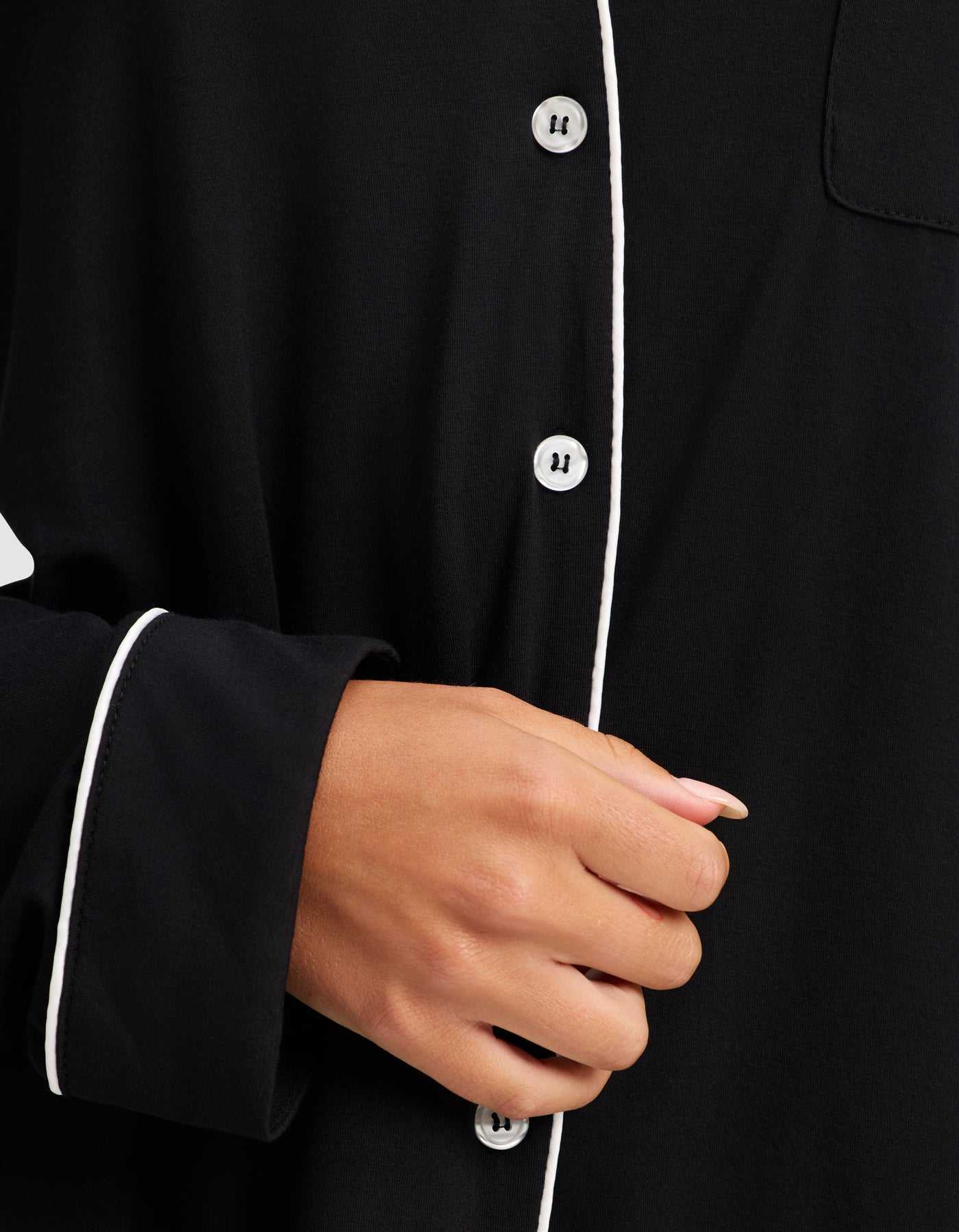A close-up shows a hand holding the front of Cozy Earth's Women's Bamboo Stretch-Knit Long Sleeve Pajama Set, featuring white piping, white buttons, and a relaxed fit. The person’s nails are neatly manicured. |Color:Black